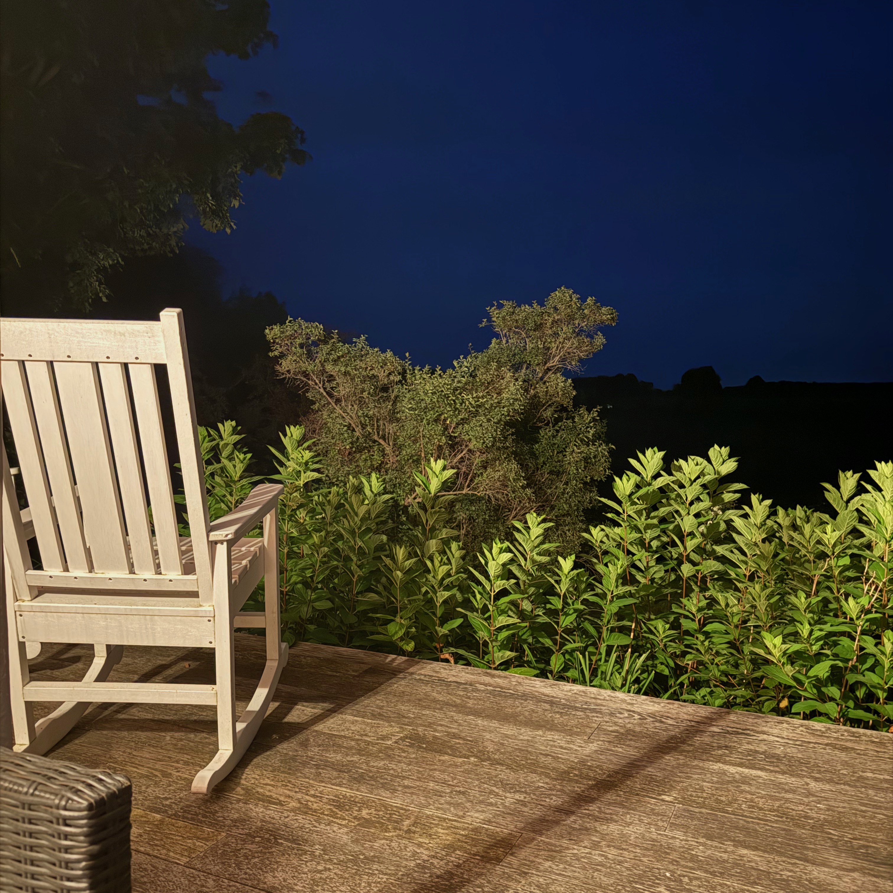 Wooden rocking chair on a deck at night, lit by a warm light, facing a row of green shrubs and a dark blue sky.