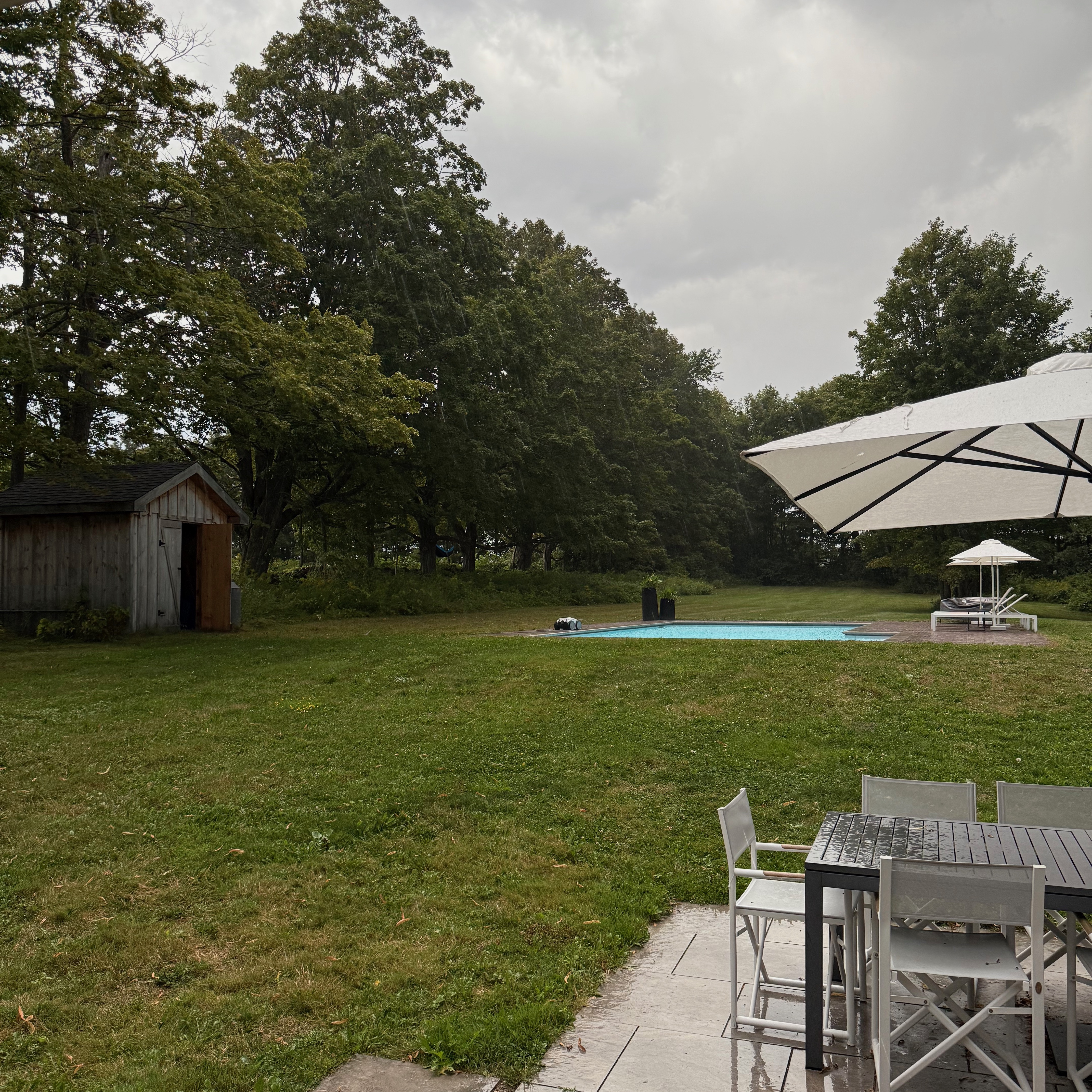 Rainy backyard scene with wet patio furniture in the foreground, a white umbrella, a swimming pool in the middle ground, and a small wooden shed near the tree line.