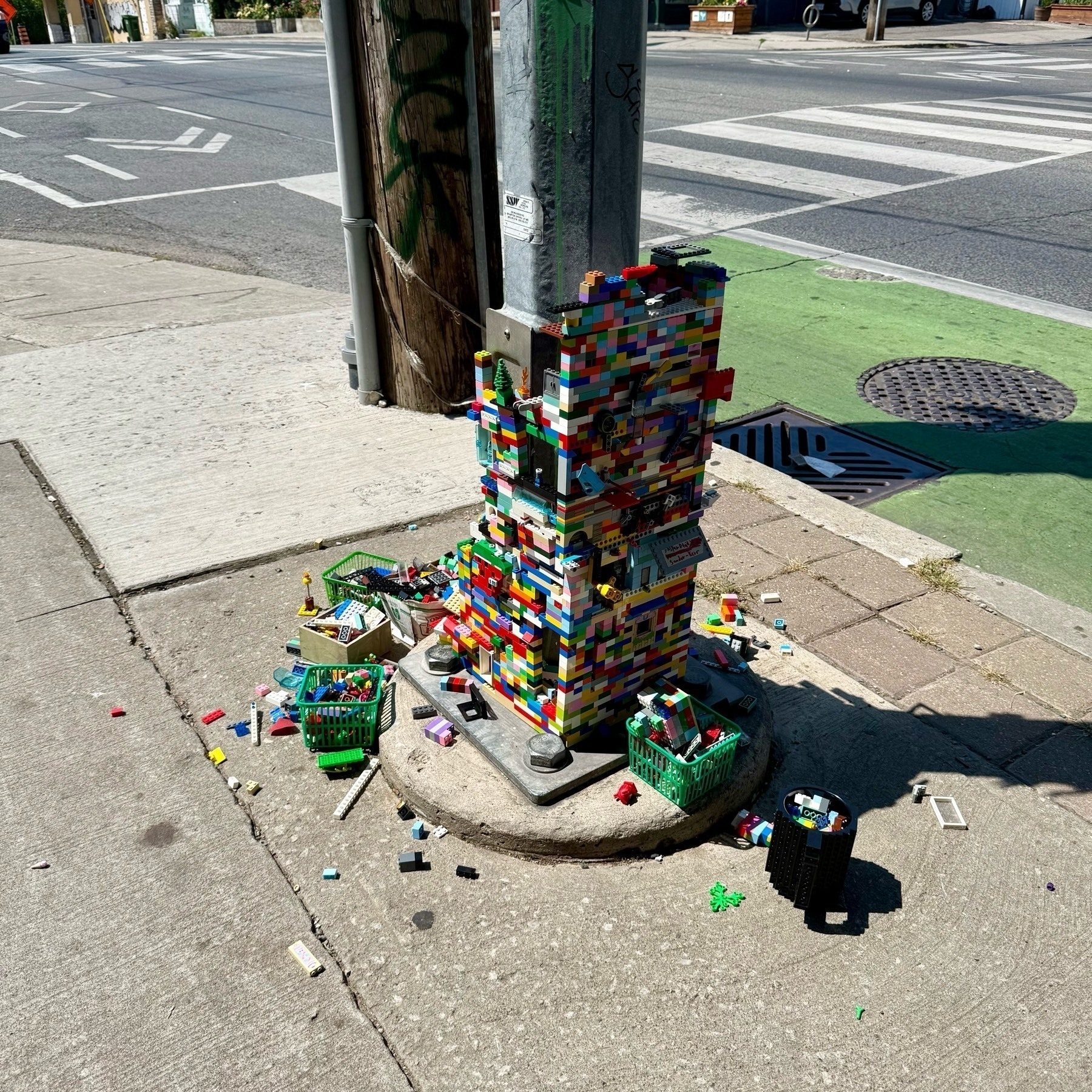 A traffic light pole base on a city sidewalk is covered in colourful LEGO bricks, forming a tall, blocky structure. Small baskets filled with loose LEGO pieces surround the base, and scattered bricks are spread across the pavement.