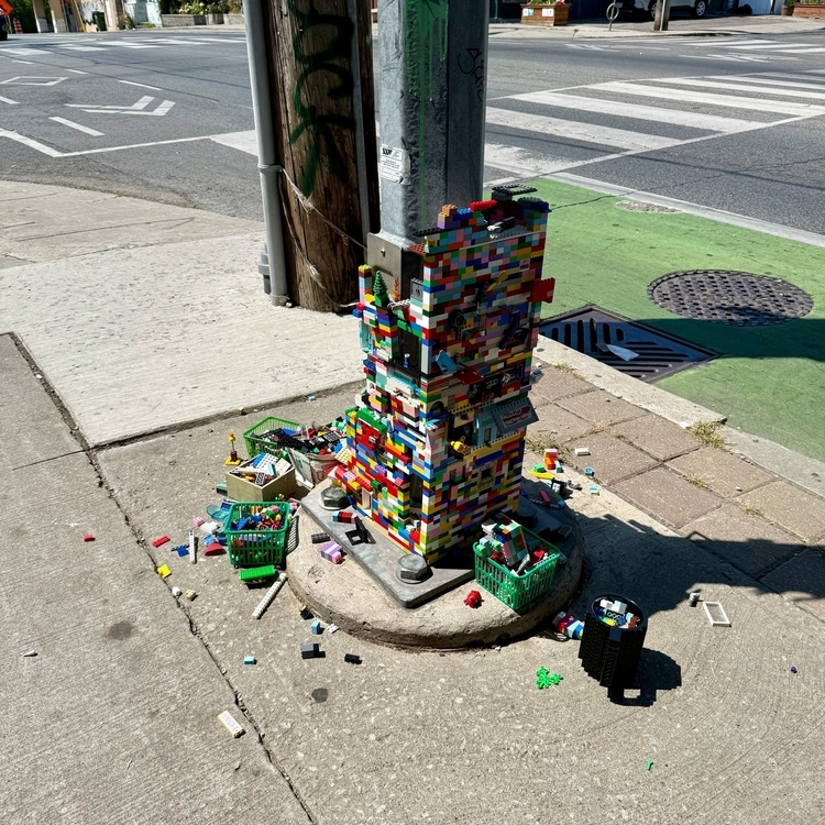A traffic light pole base on a city sidewalk is covered in colourful LEGO bricks, forming a tall, blocky structure. Small baskets filled with loose LEGO pieces surround the base, and scattered bricks are spread across the pavement.
