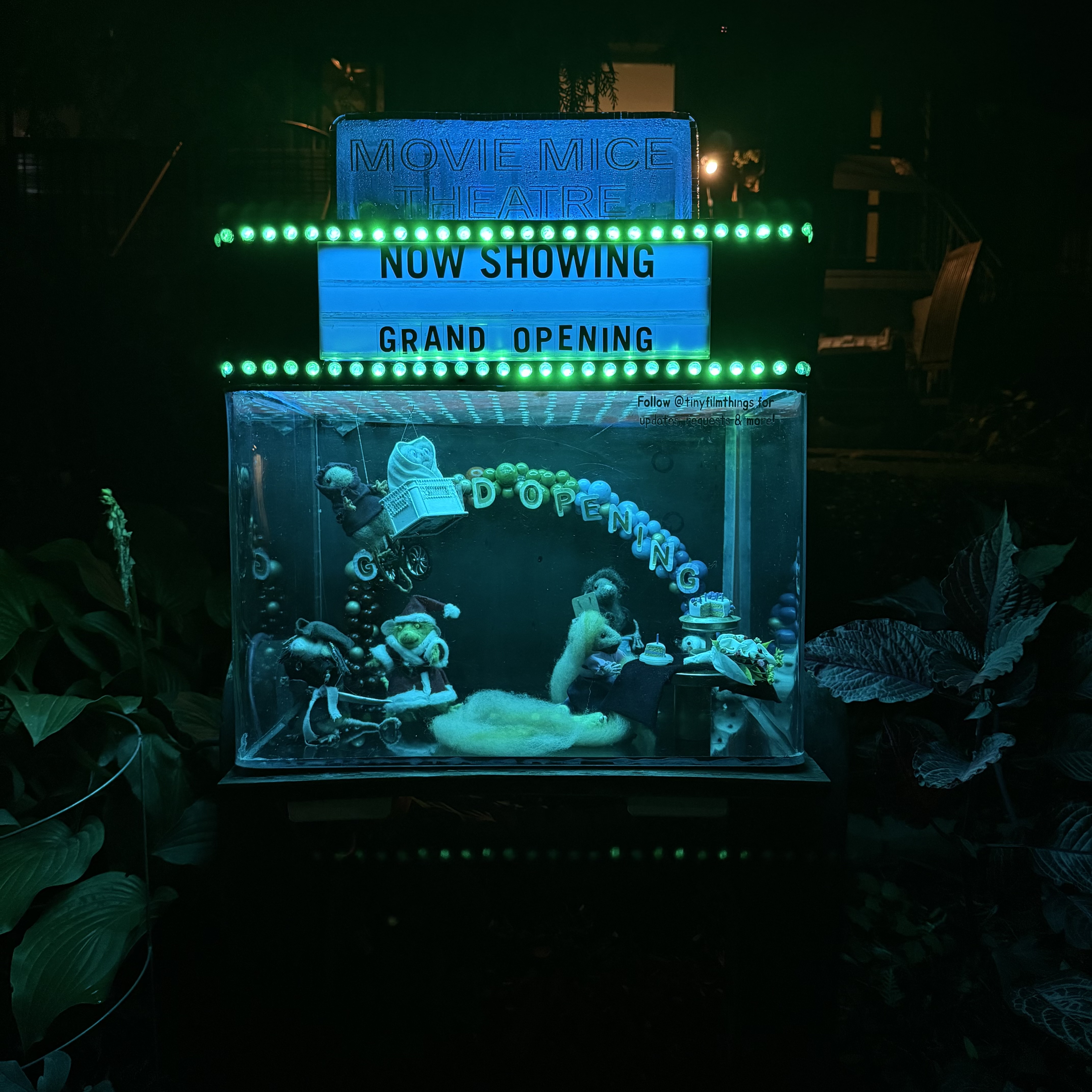 A whimsical miniature diorama of a “Movie Mice Theatre” is lit with glowing green lights at night. A marquee above reads “NOW SHOWING — GRAND OPENING.” Inside a clear display box below, tiny mouse figurines are posed at a grand opening celebration, complete with streamers, balloons, and a “Grand Opening” banner. One mouse sits at a small table with a cake, another pushes a baby carriage, and others are dressed up as if attending a party. The whole scene is nestled among garden plants, giving it a magical, hidden-world feel.