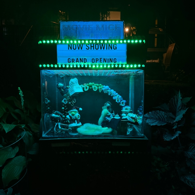 A whimsical miniature diorama of a “Movie Mice Theatre” is lit with glowing green lights at night. A marquee above reads “NOW SHOWING — GRAND OPENING.” Inside a clear display box below, tiny mouse figurines are posed at a grand opening celebration, complete with streamers, balloons, and a “Grand Opening” banner. One mouse sits at a small table with a cake, another pushes a baby carriage, and others are dressed up as if attending a party. The whole scene is nestled among garden plants, giving it a magical, hidden-world feel.