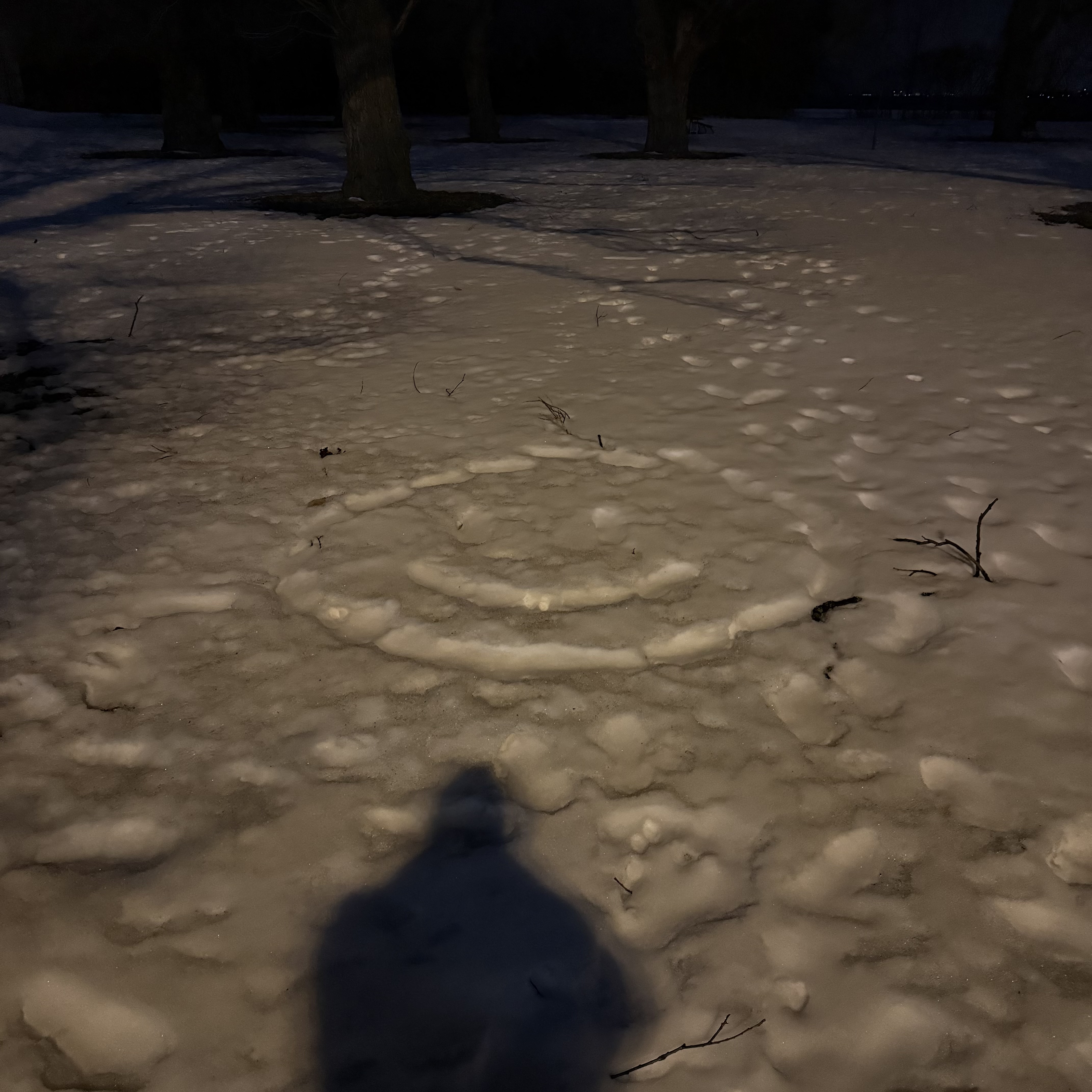 A smiley face drawn in snow on a park ground at night, lit by warm amber streetlight. The photographer's shadow stretches into the foreground.