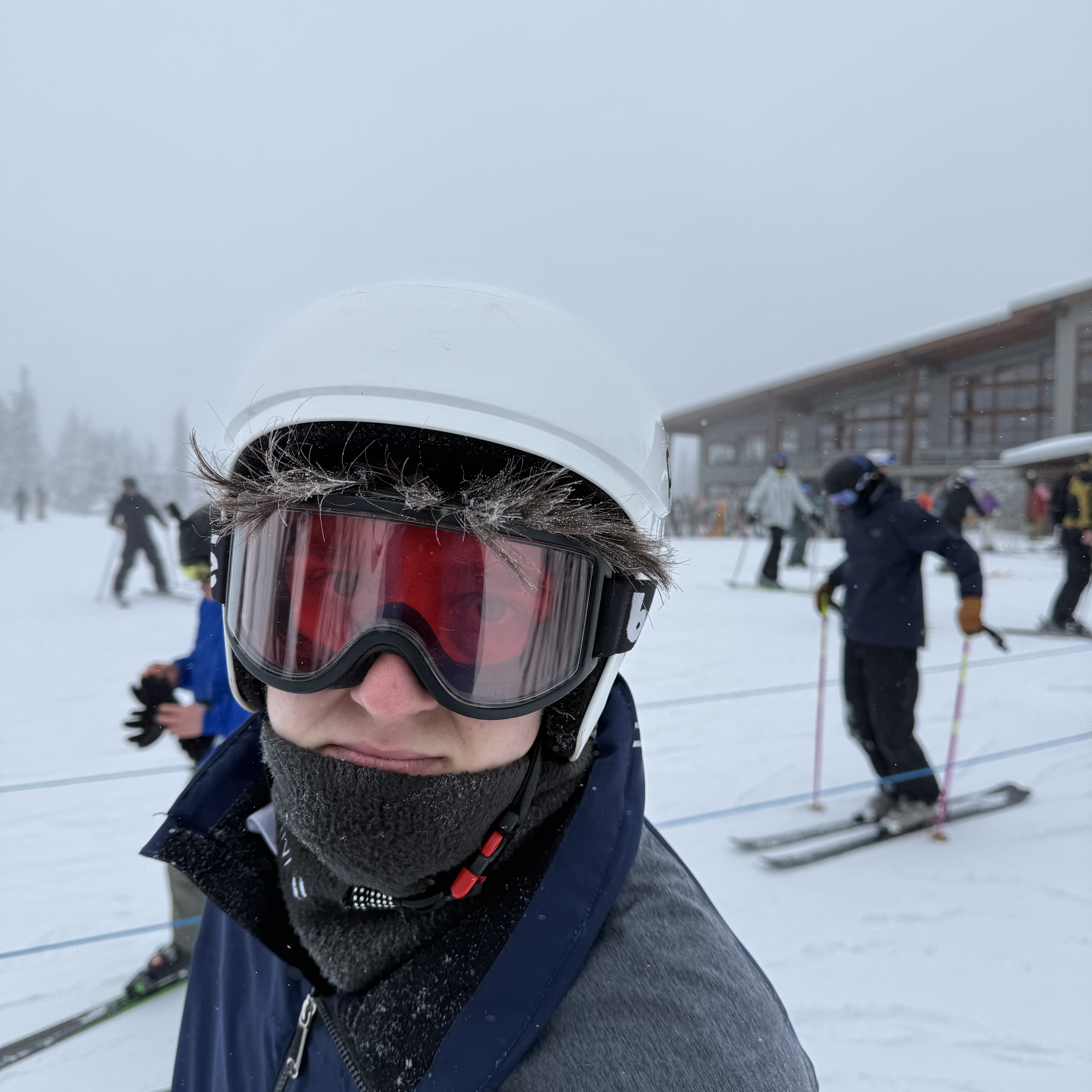 Hayden in ski gear — white helmet, red-lens goggles, and a black neck gaiter — at the base of a ski resort on an overcast, snowy day. Other skiers and a lodge building are visible in the background.