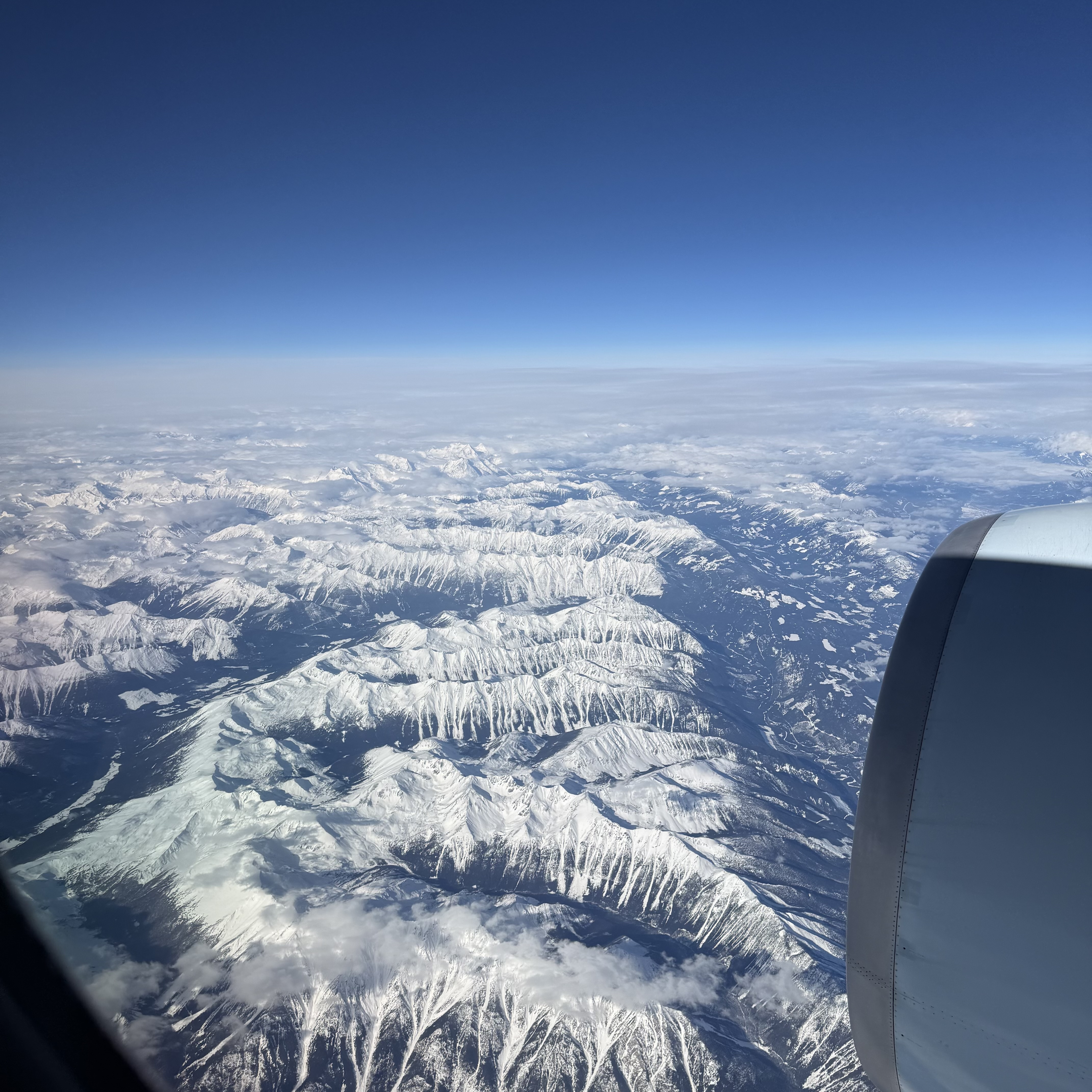 Aerial view of snow-covered mountain ranges stretching to the horizon under a deep blue sky, with a jet engine visible in the lower right corner and patches of cloud drifting between the peaks.