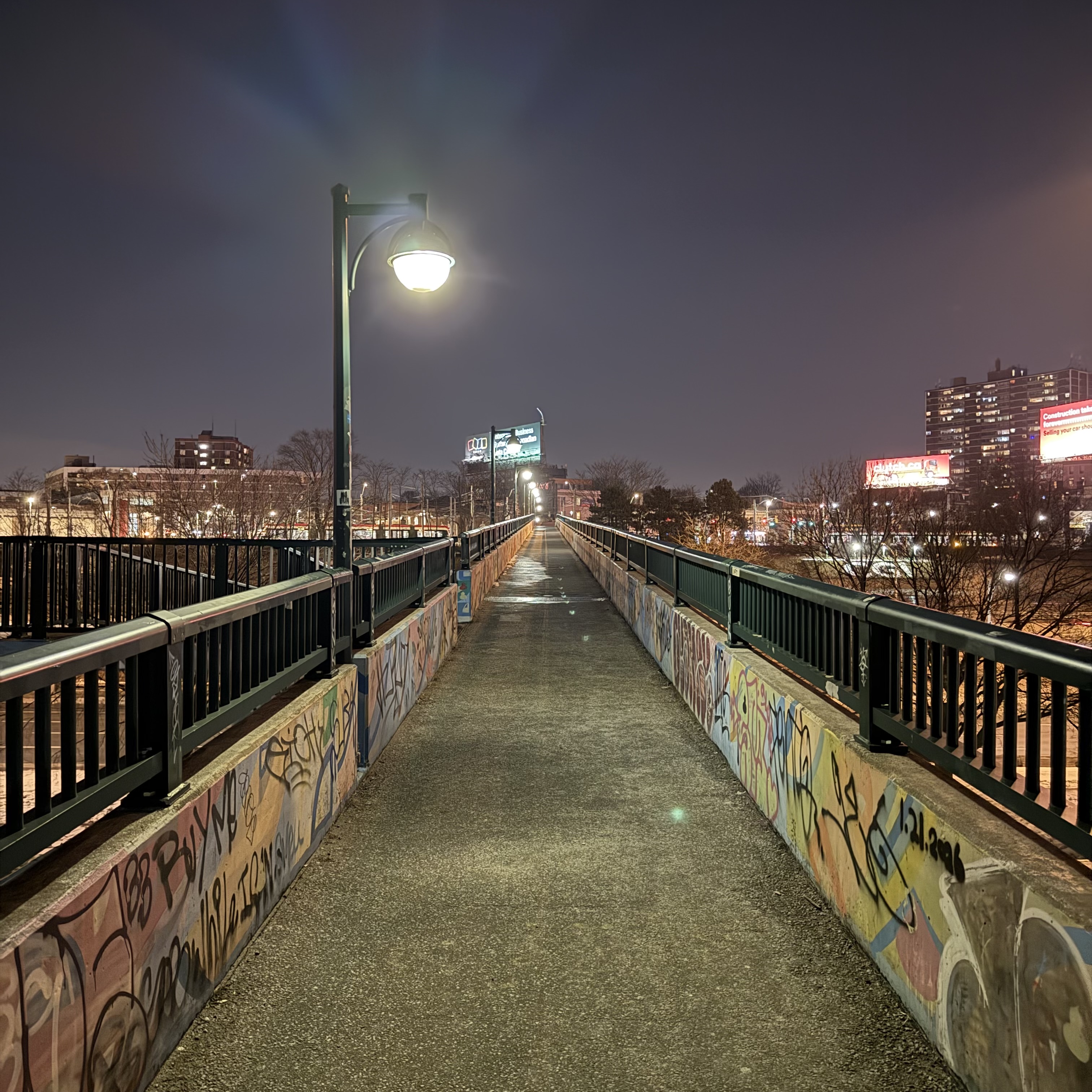 A pedestrian bridge at night, its concrete barriers covered in colourful graffiti, stretching toward a distant city skyline lit with signs and streetlights.