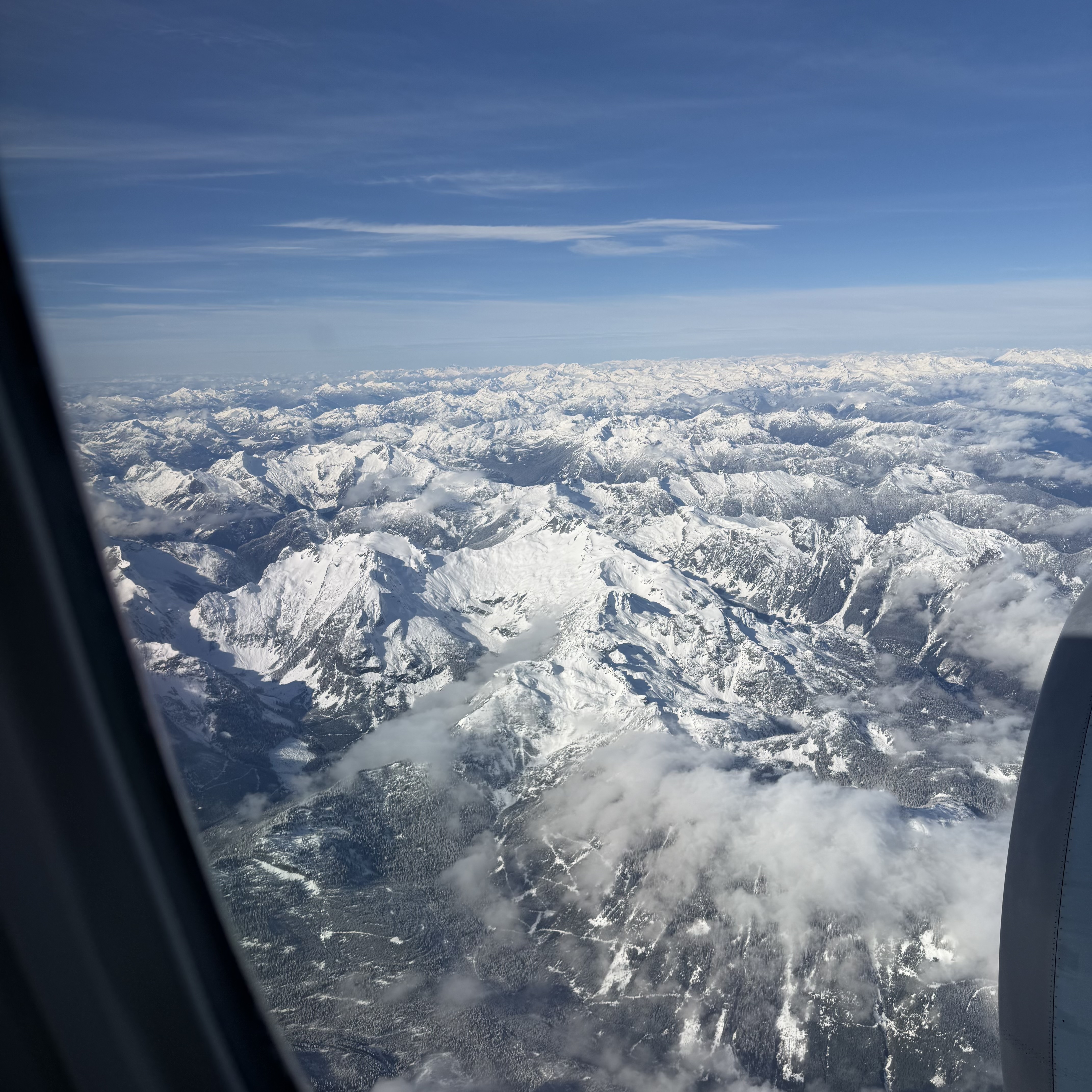 Snow-dusted mountain ranges seen through an airplane window, with dark evergreen forest visible in the valleys below and billowing white clouds nestled between the ridgelines.