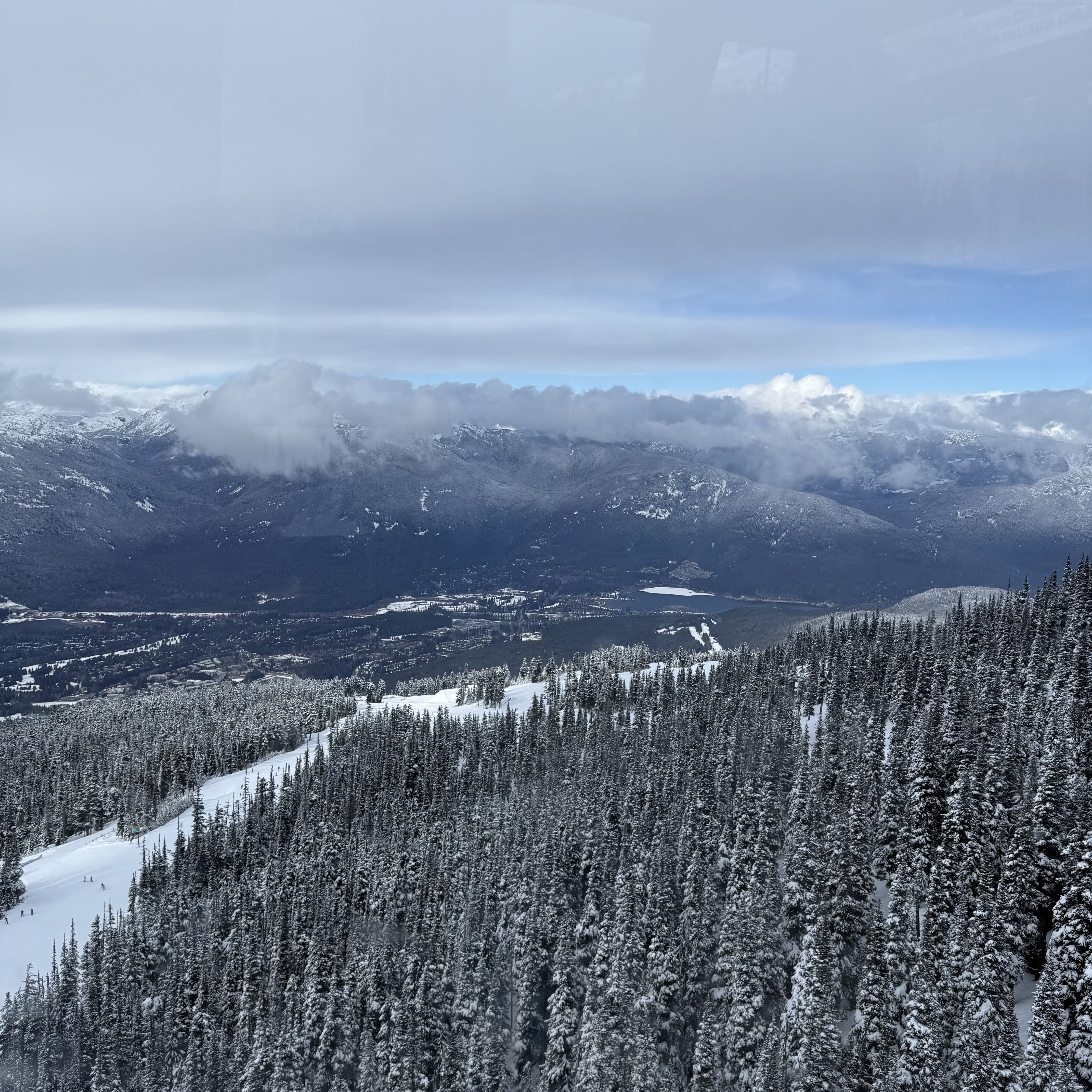 A sweeping view from high on Whistler Mountain looking down over snow-laden pine trees and ski runs into the valley below, with Whistler village visible in the distance under a partly cloudy blue sky.