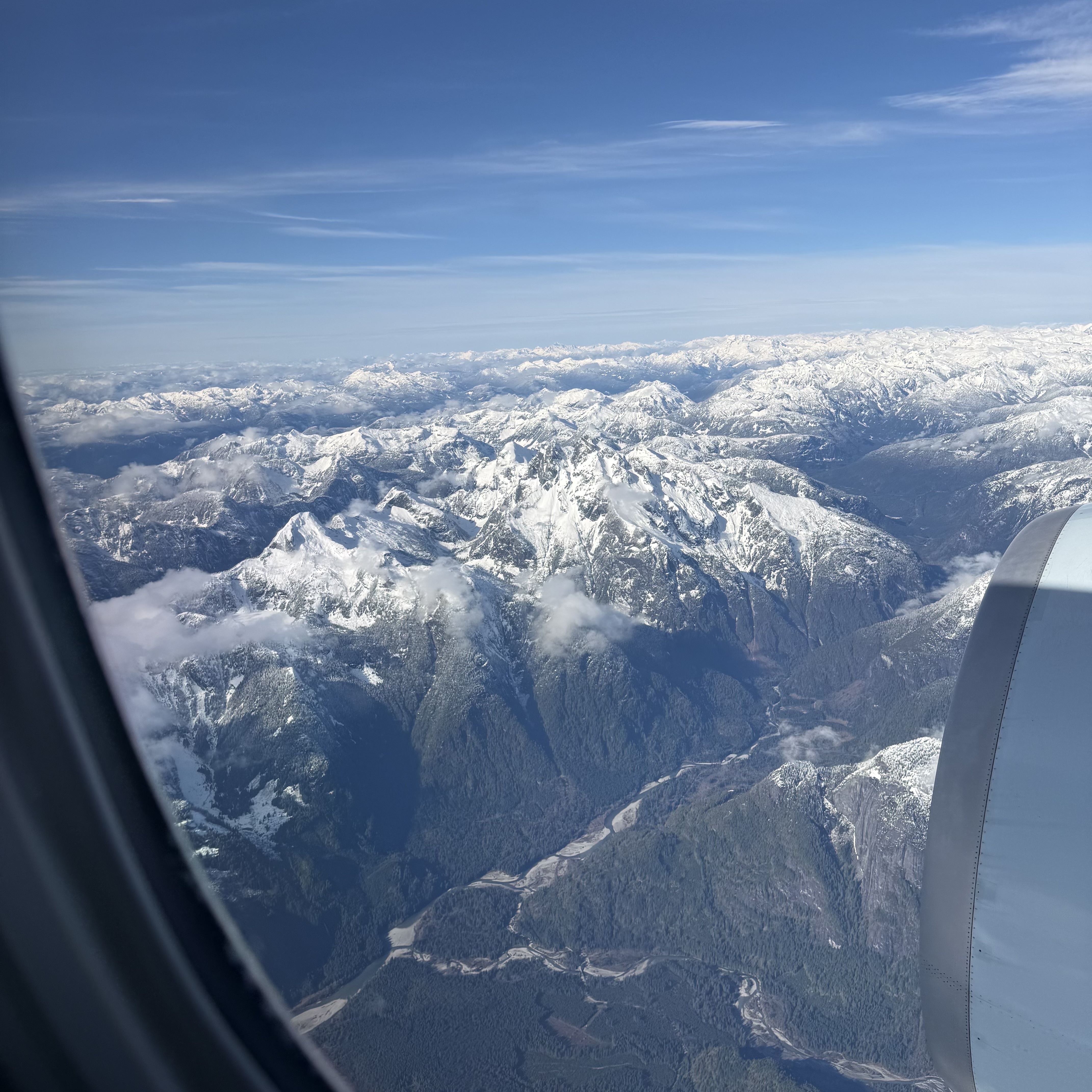 A jagged cluster of snow-capped peaks framed by an airplane window, with a river valley and dense forest cutting through the lower terrain and the engine nacelle visible at the right edge of the frame.