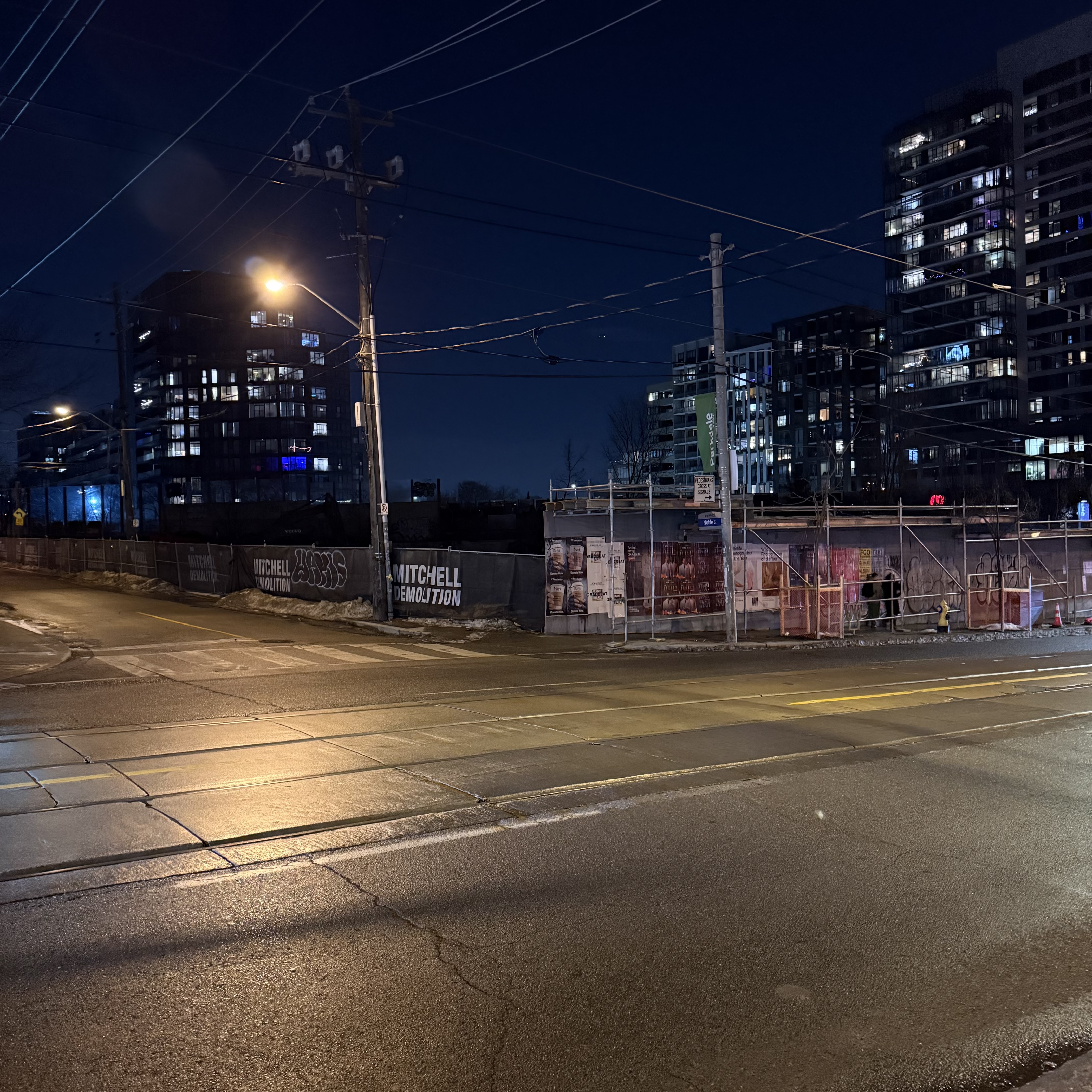 A quiet city intersection at night. A construction hoarding reads "Mitchell Demolition" along a fenced-off empty lot, with condos glowing in the background.