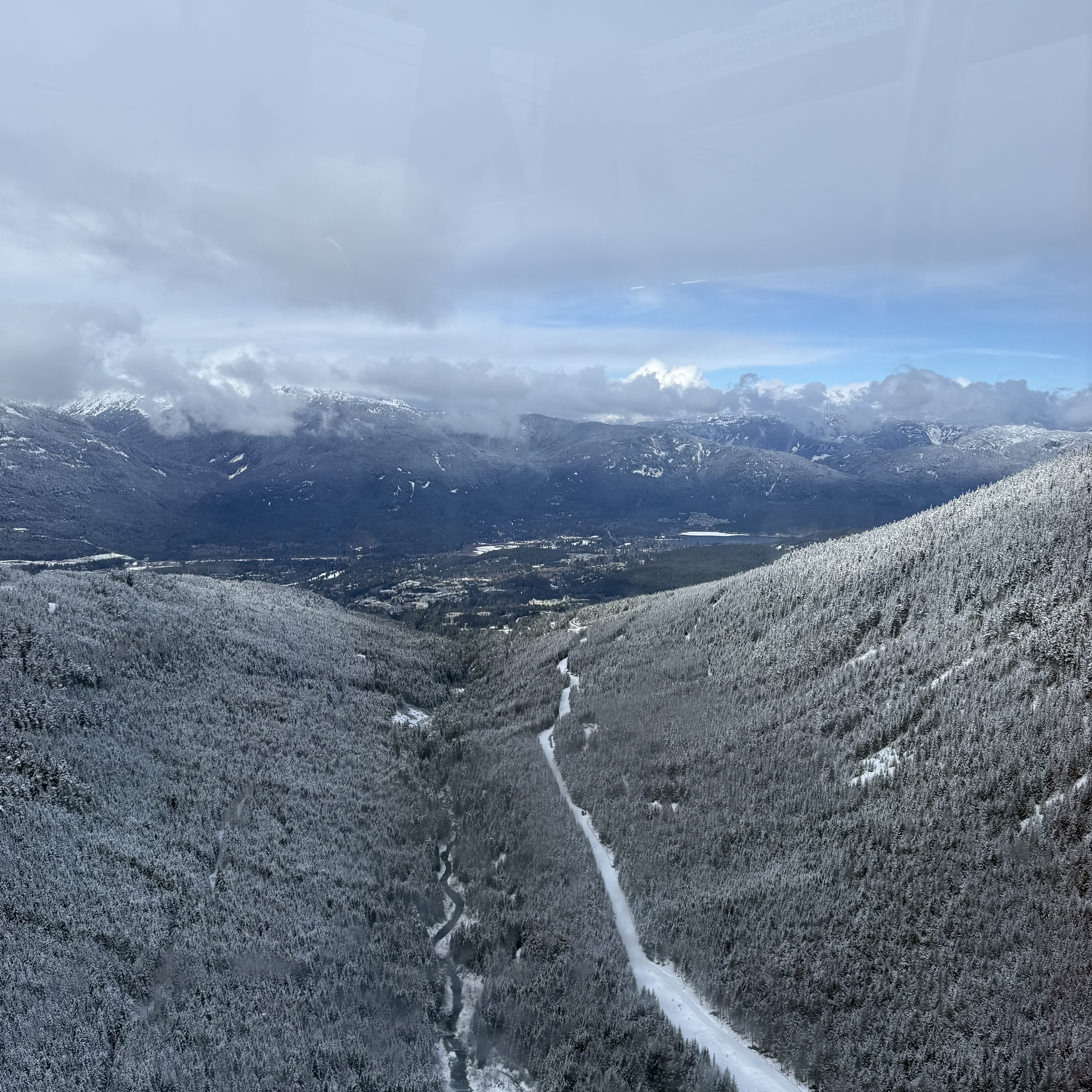 Looking down a wide mountain valley from a gondola, with snow-covered trees blanketing both hillsides, a ski run cutting a straight line through the centre, and the Whistler valley floor visible far below.