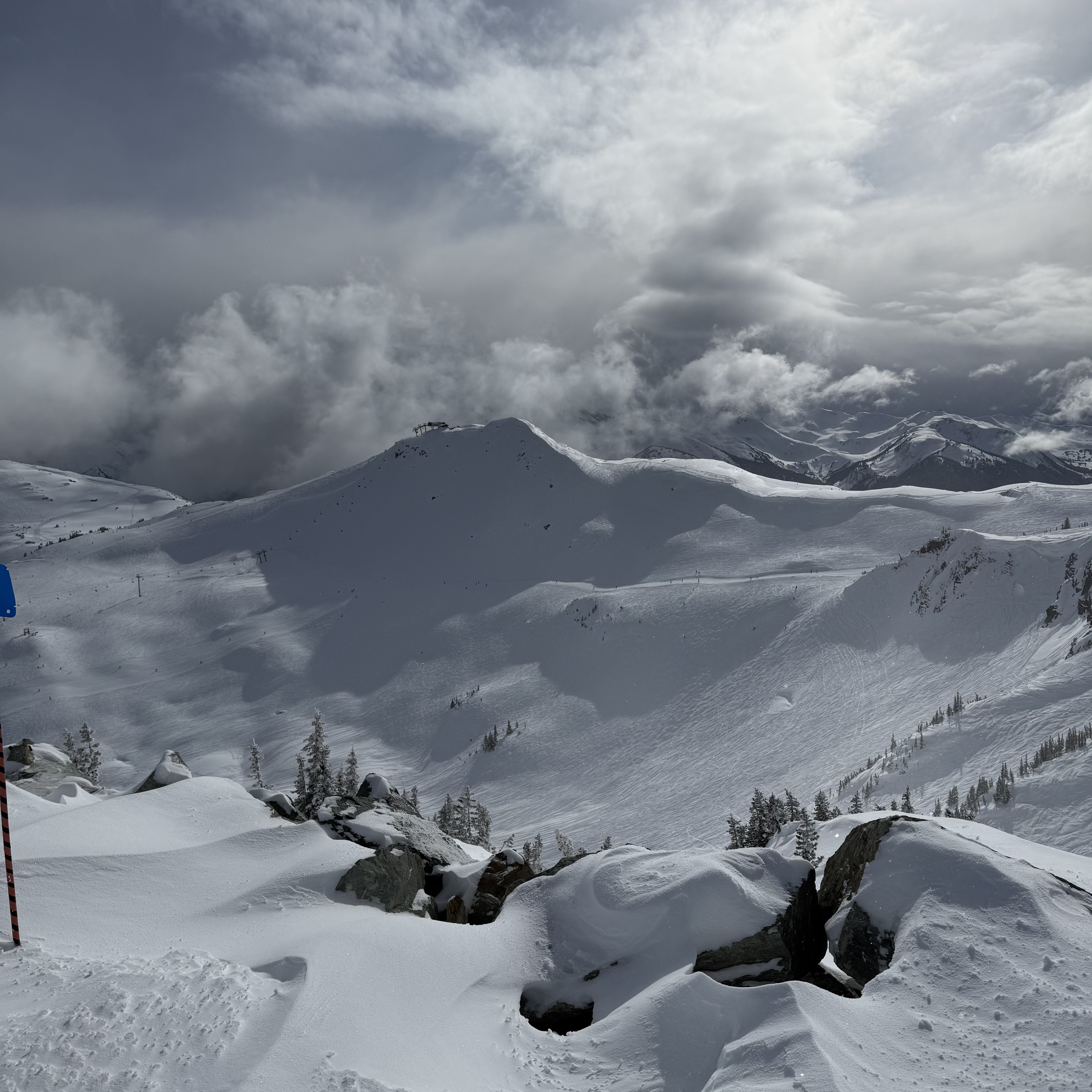 High alpine terrain at Whistler with dramatic storm clouds rolling over steep, snow-covered peaks. Rocky outcroppings are buried under deep snow in the foreground, and a blue trail marker is barely visible at the left edge.