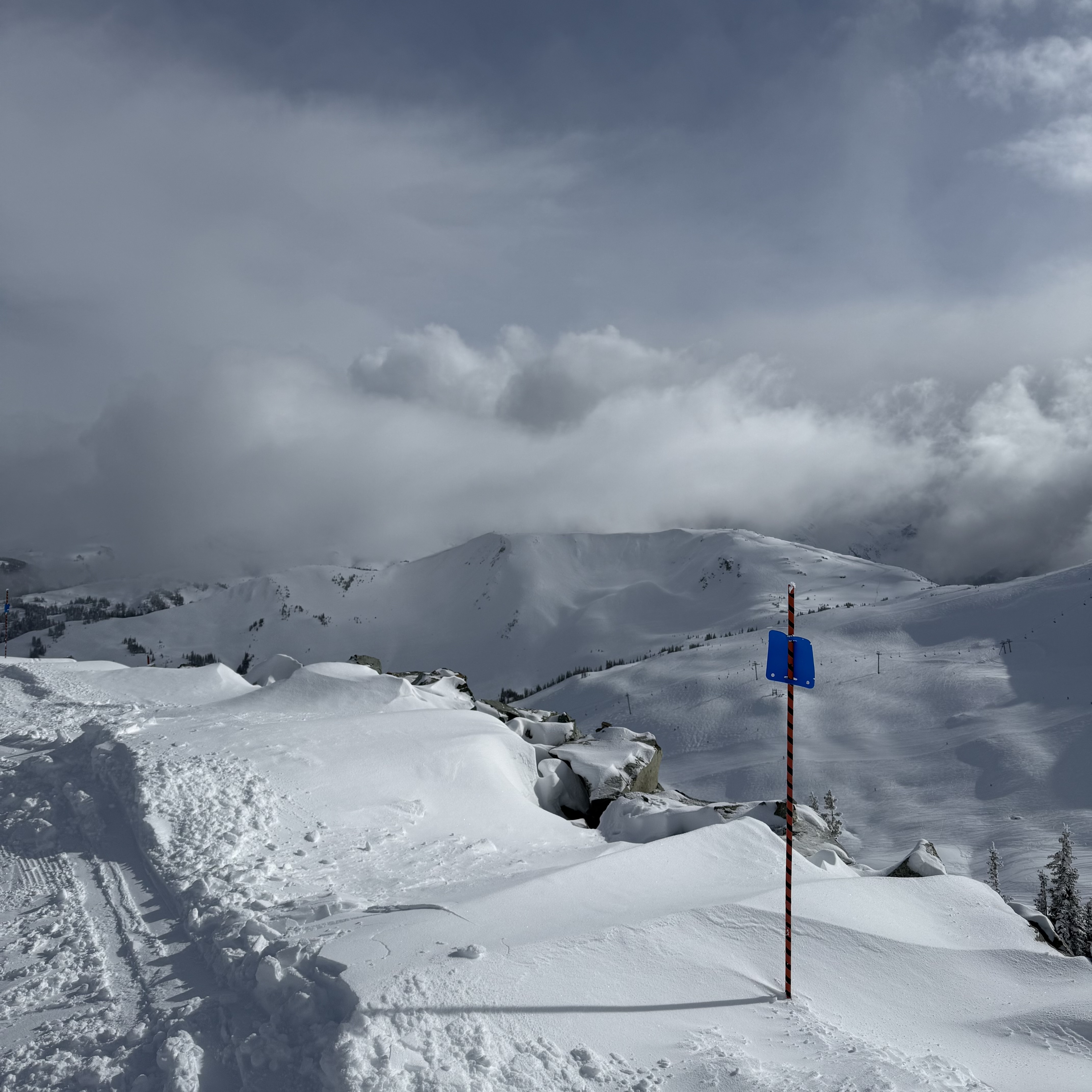 A blue diamond ski trail marker on a red and white pole stands on a snowy alpine ridge at Whistler, with wind-sculpted snowdrifts over rocks in the foreground and overcast mountain peaks stretching into the distance.