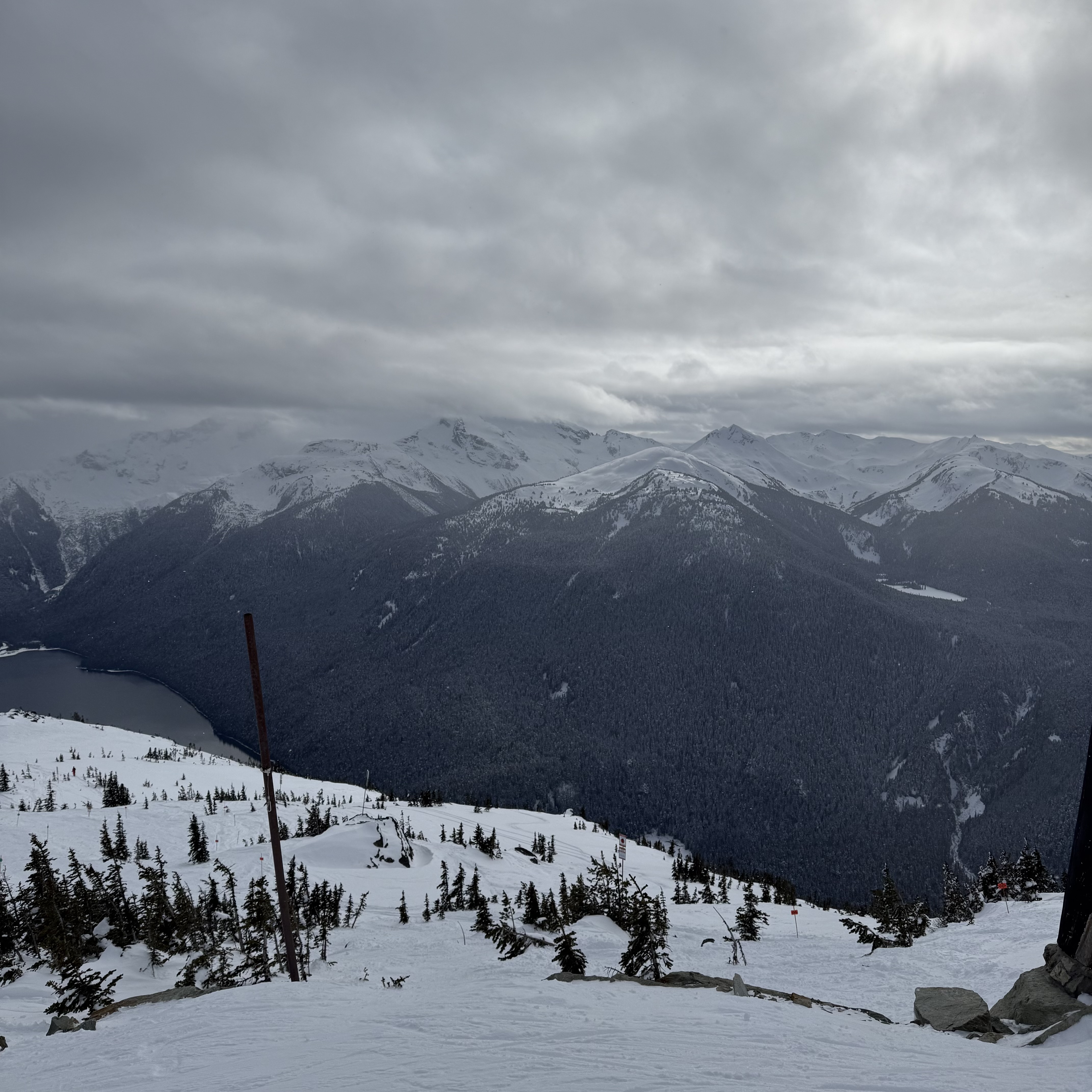 Looking out from a high ridge on Blackcomb Mountain over a frozen lake nestled in the valley far below, surrounded by dark forested peaks under a heavy overcast sky. Ski runs and scattered pines dot the slope in the foreground.