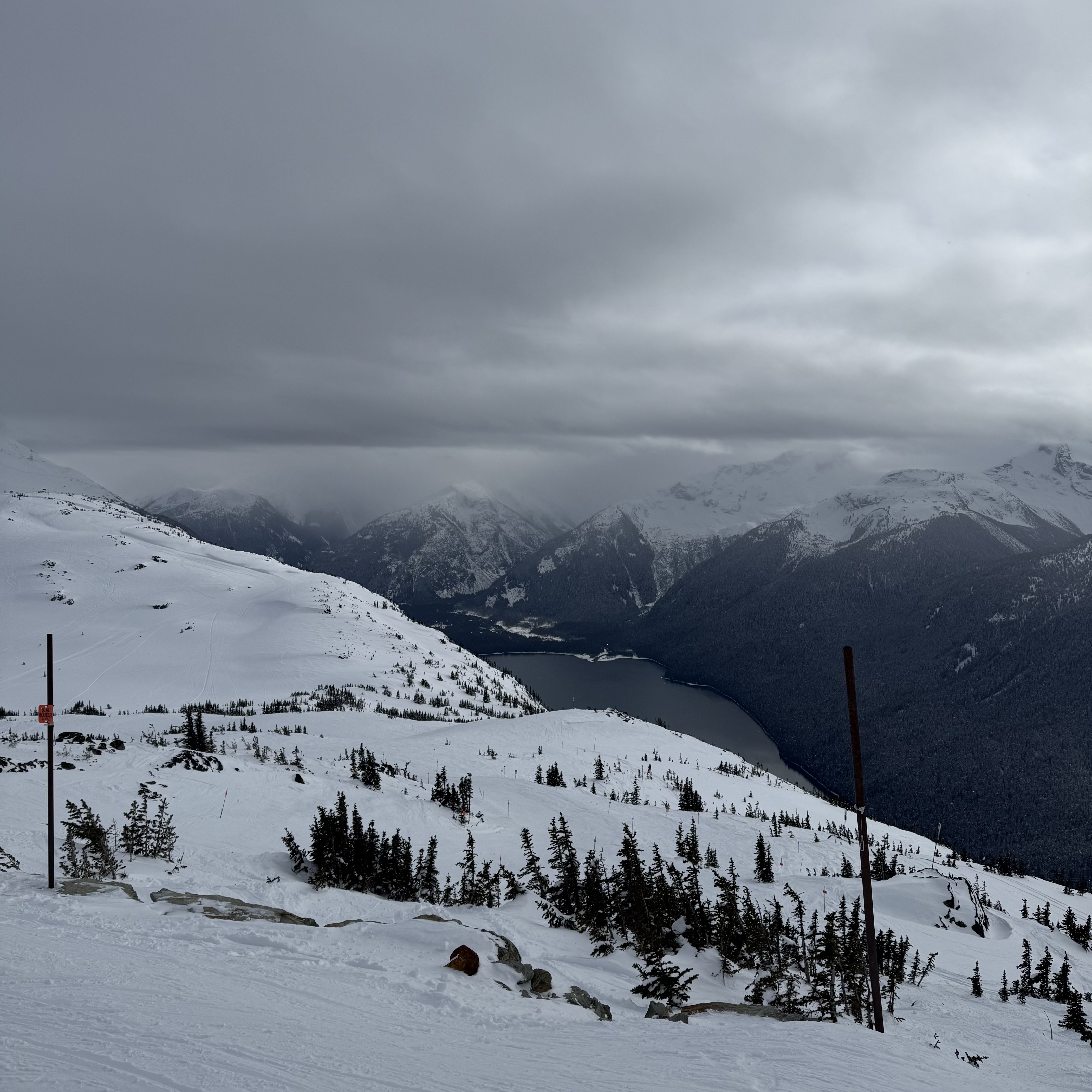 A long, narrow lake — Green Lake — winds through the valley below, framed by snow-covered slopes, scattered pines, and trail markers. Moody grey clouds drape over the mountain ridgeline in the background.