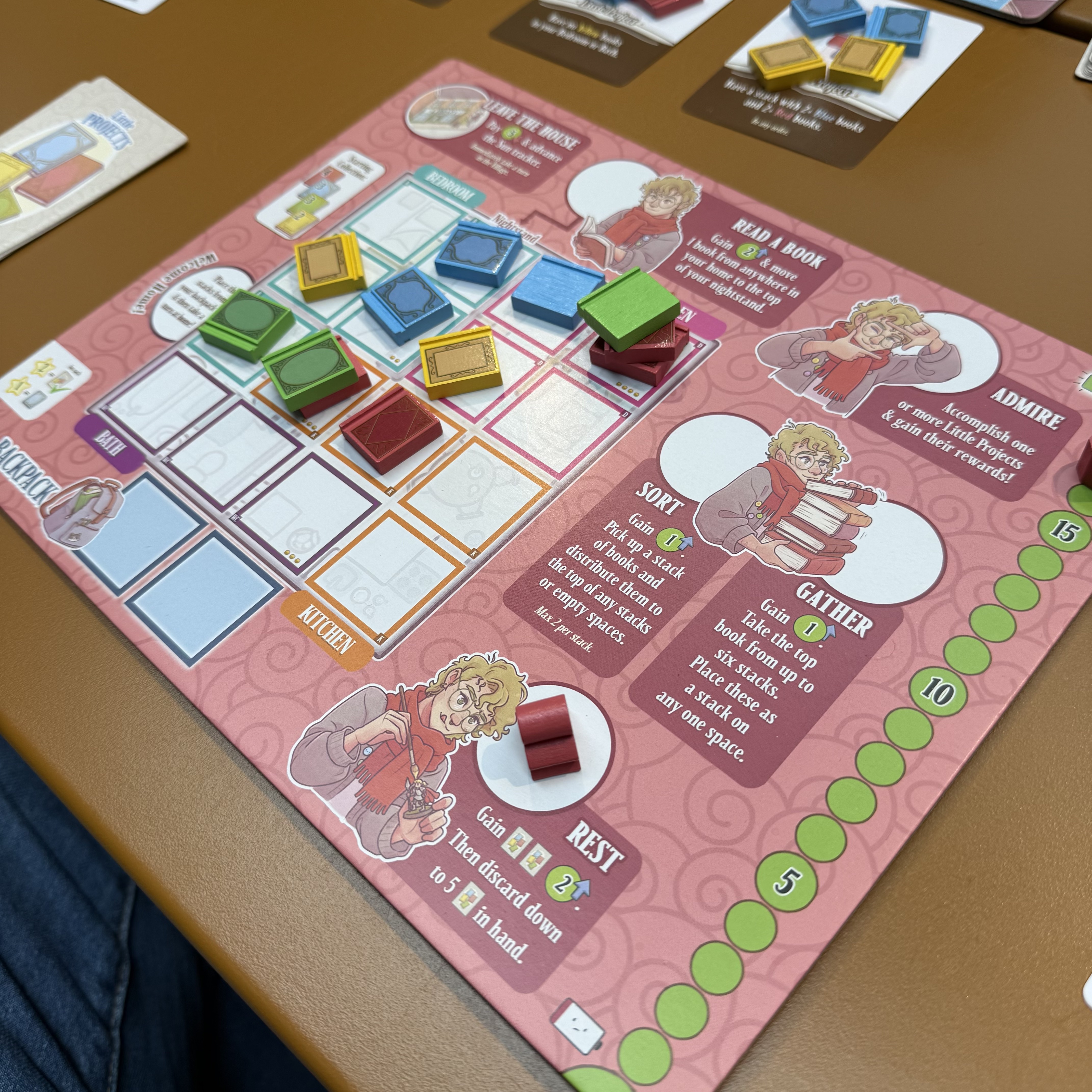 A pink game board mid-play, action spaces labeled Rest, Gather, Sort, Read a Book, and Admire. Colourful wooden book tokens in green, blue, yellow, and red are spread across a room grid: Bedroom, Bath, Kitchen, Backpack.