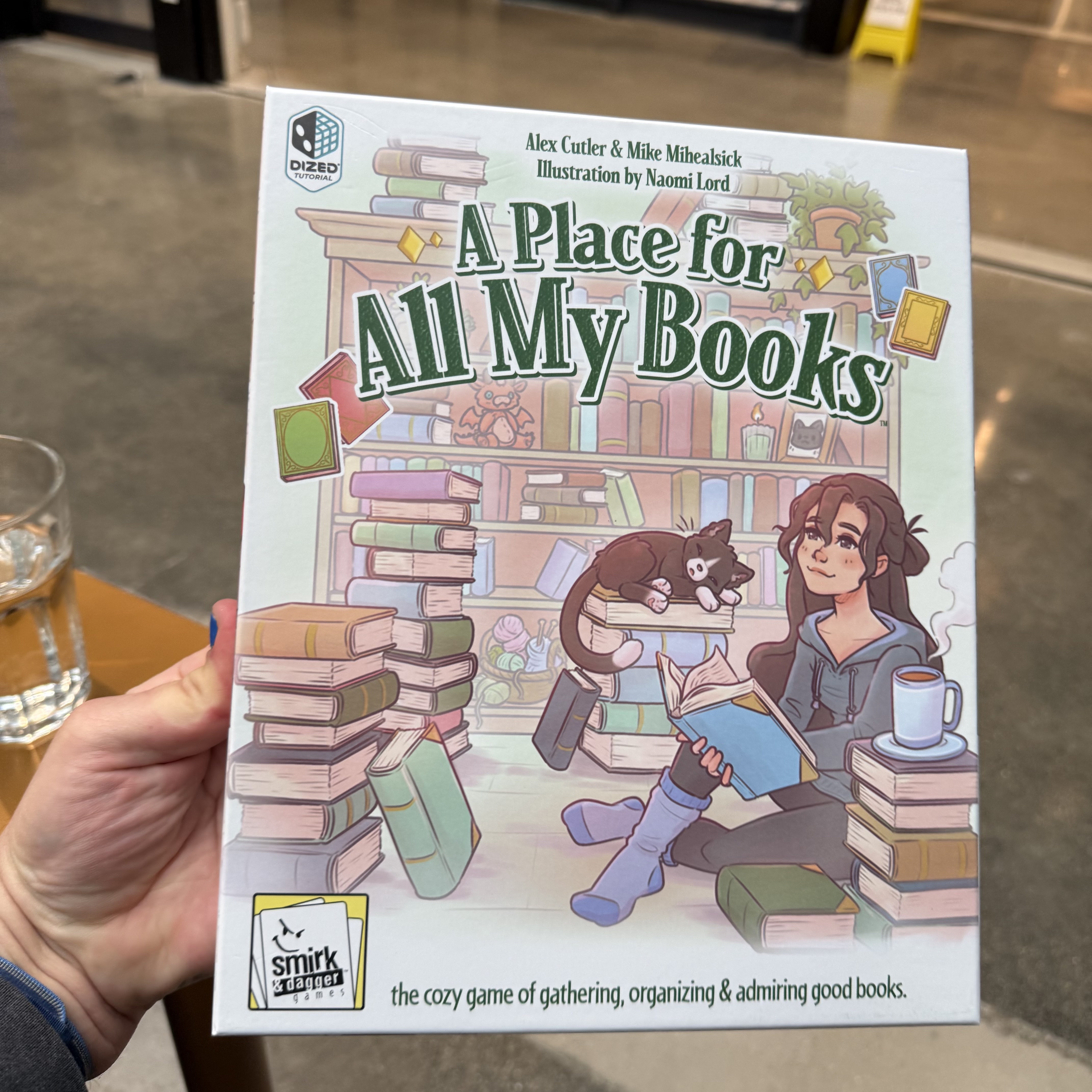 A hand holds the box for A Place for All My Books, a Smirk & Dagger game illustrated by Naomi Lord. The cover shows a young woman reading amid towering book stacks, a black cat draped across her lap, a steaming mug nearby.