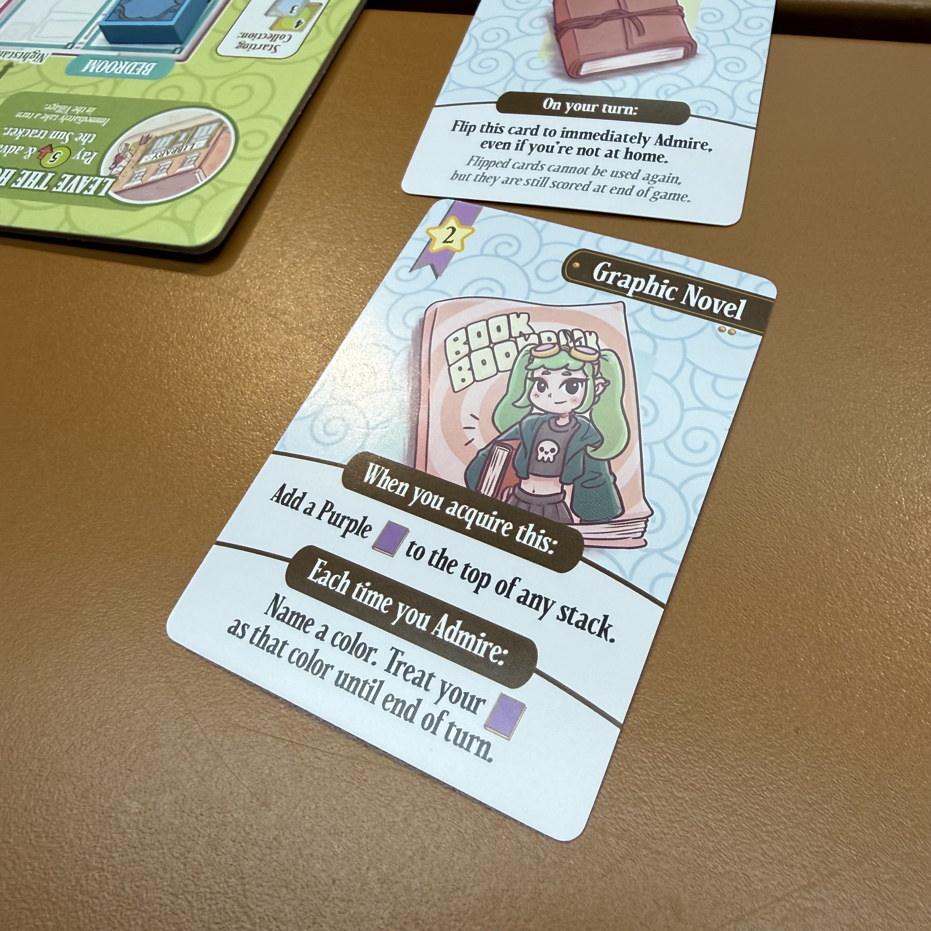 A Graphic Novel card featuring an anime-style character with long green hair and a skull tee. Acquire effect: add a purple book to any stack. Admire effect: name a color, treat your purple book as that color until end of turn.