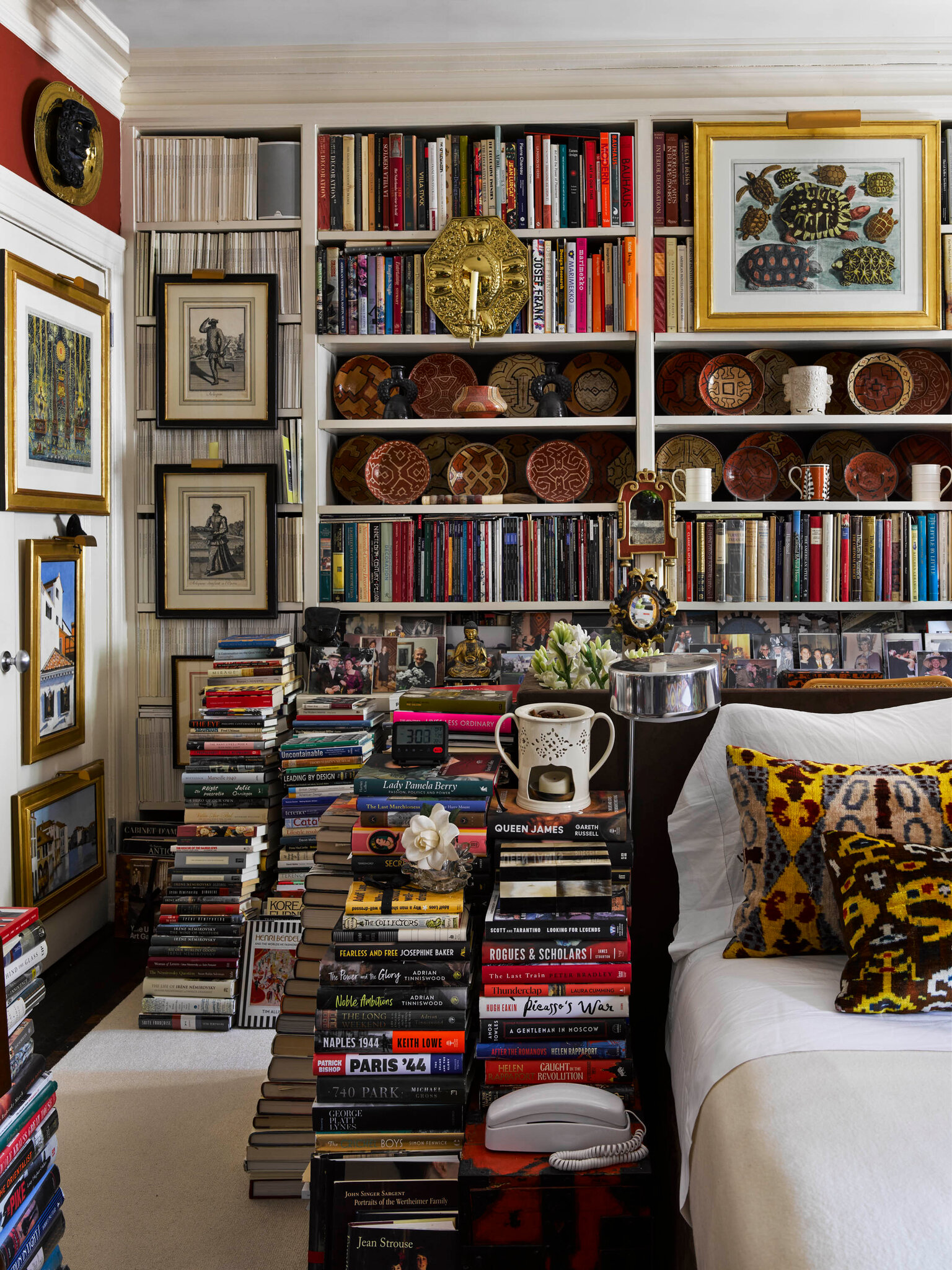 A bedroom corner overflowing with tall stacks of books on the floor and used as nightstands. Built-in shelves behind the bed are packed with books, pottery, and framed art. A white bed with patterned pillows sits beside the vertical towers of books, alongside small objects like a lamp, flowers, and a vintage telephone.