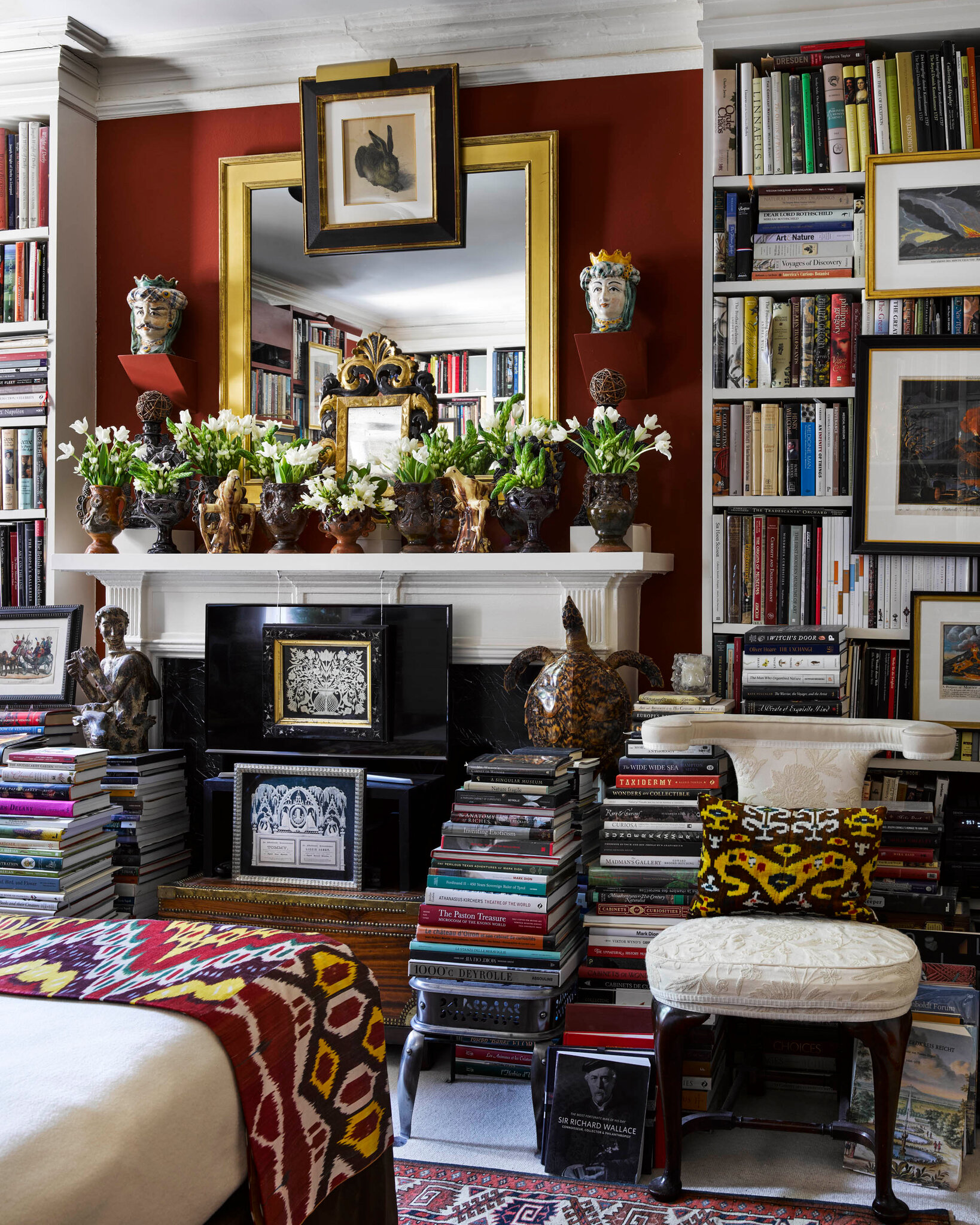 Another view of the bedroom, showing a fireplace mantel crowded with decorative vases and fresh flowers, backed by a large mirror and framed art. Bookshelves overflow with books on either side, and more floor-to-ceiling stacks rest around a pair of upholstered chairs. The room feels maximalist, warm, and deeply curated.