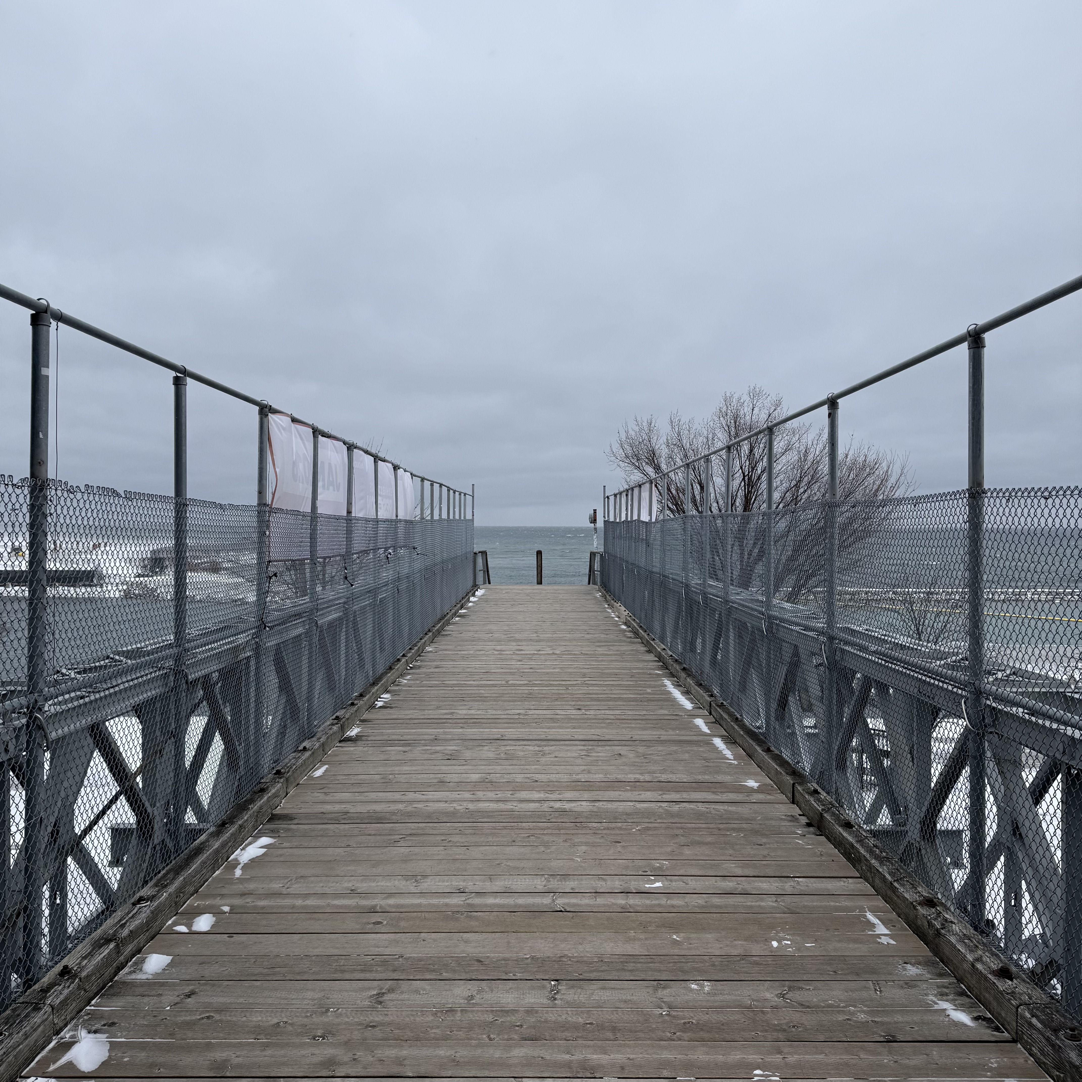 A wooden walkway fenced with chain-link leading straight out toward the lake beneath an overcast sky.