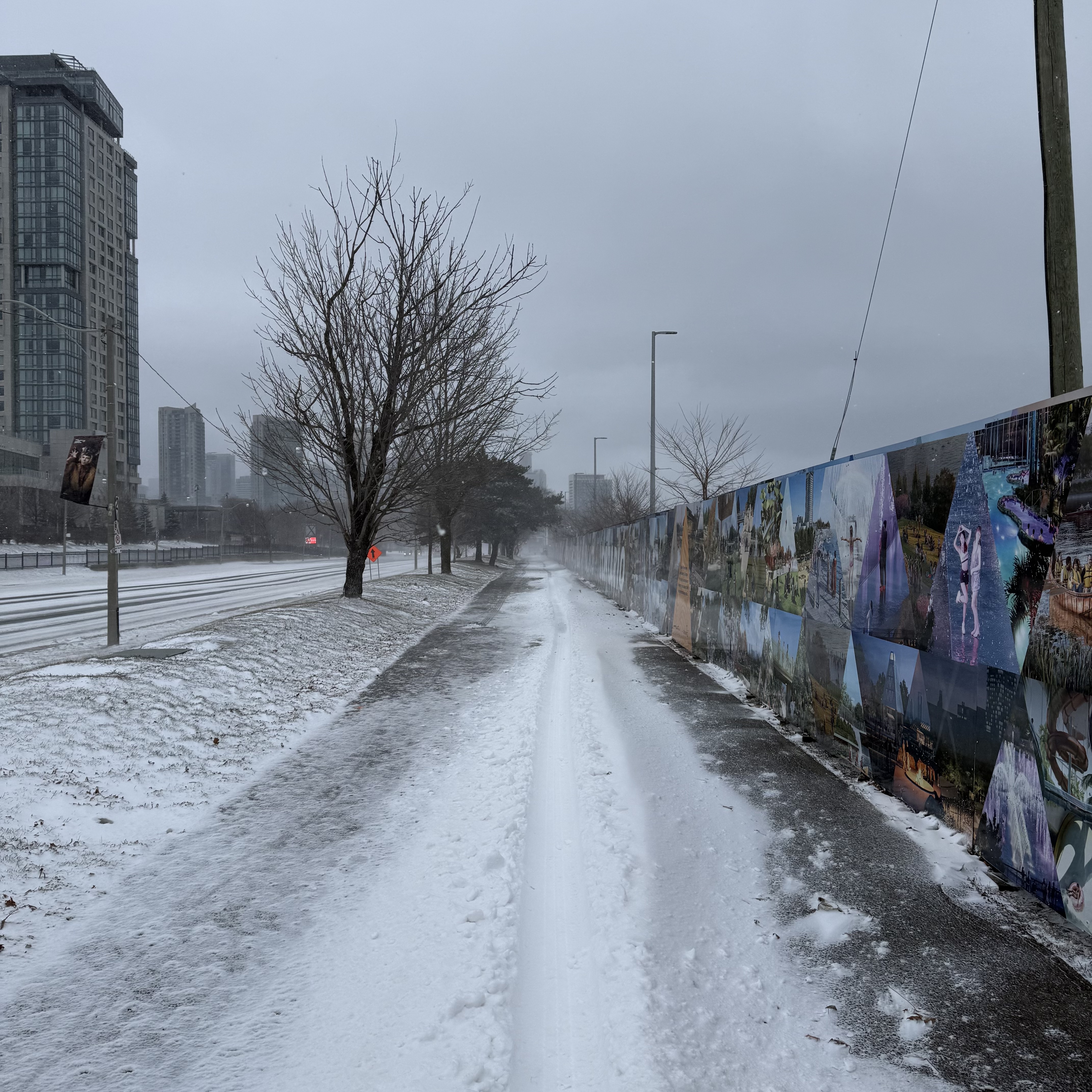 A windblown, snow-dusted sidewalk beside a long mural-covered construction fence, with high-rises fading into the distance.