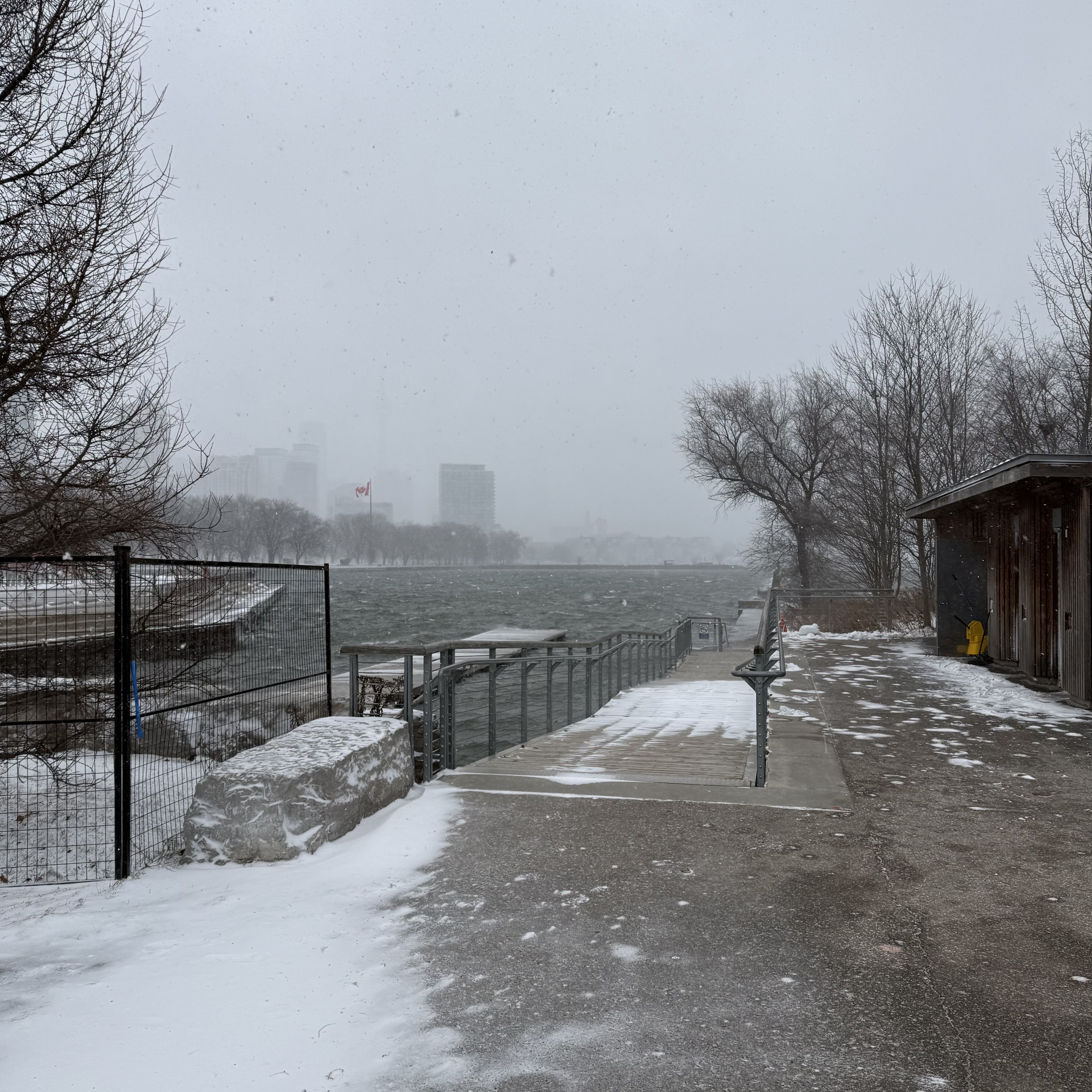 A snowy waterfront ramp with metal railings descending to choppy water, the skyline barely visible through snowfall.