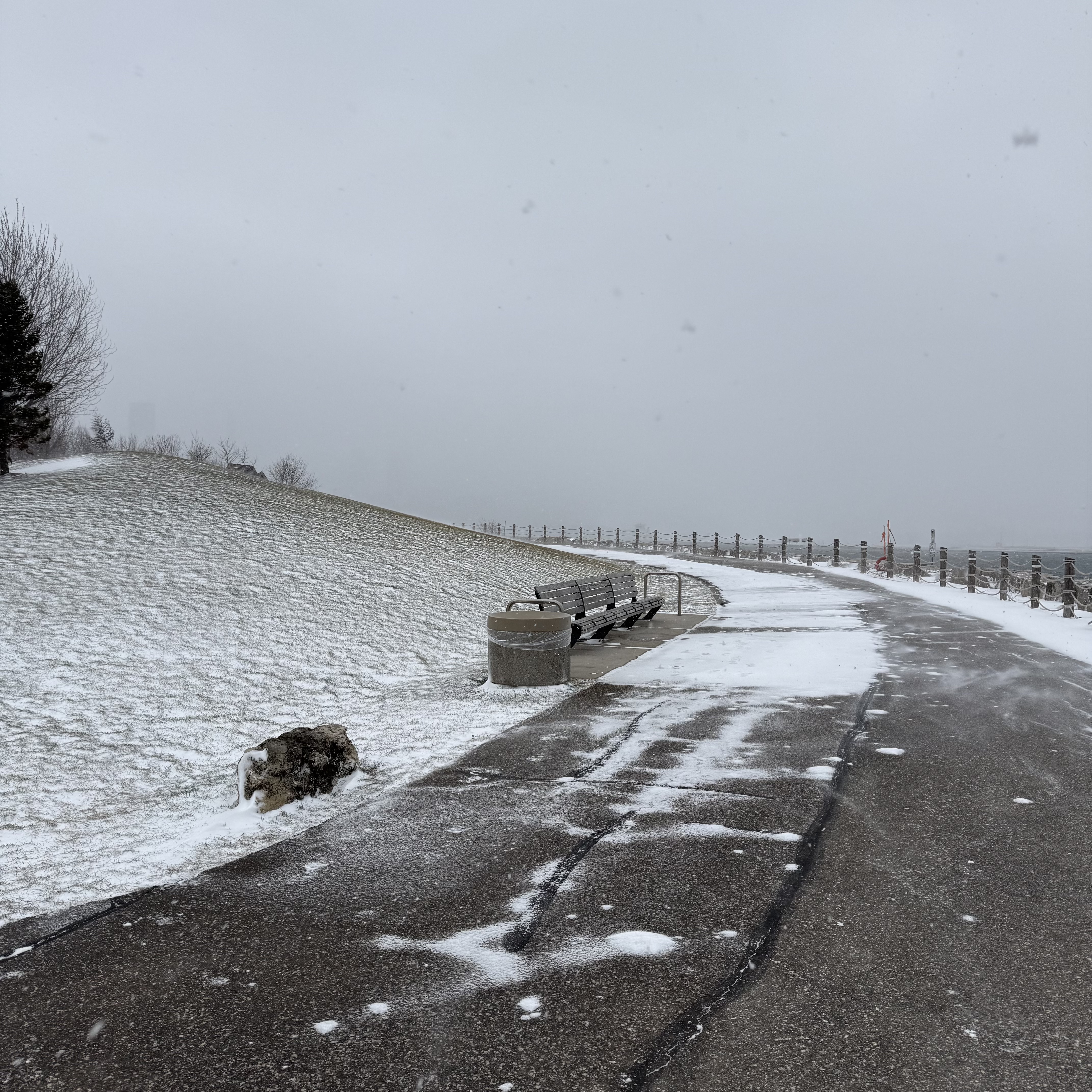 A curving lakeside path with a bench and trash can, thin snow skittering across the pavement.