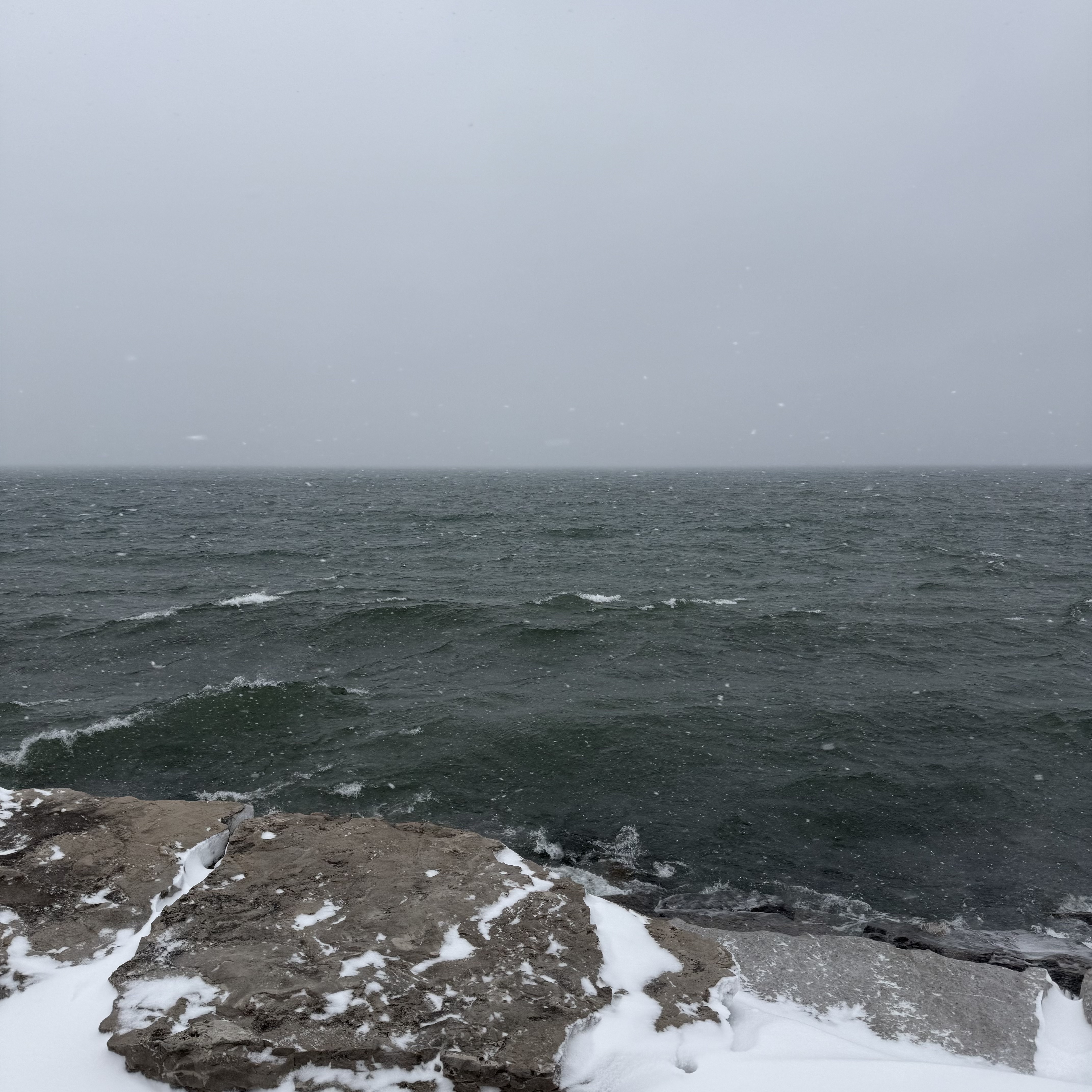 Dark green lake waves rolling in a snowstorm, viewed from a rocky, snow-covered shoreline.