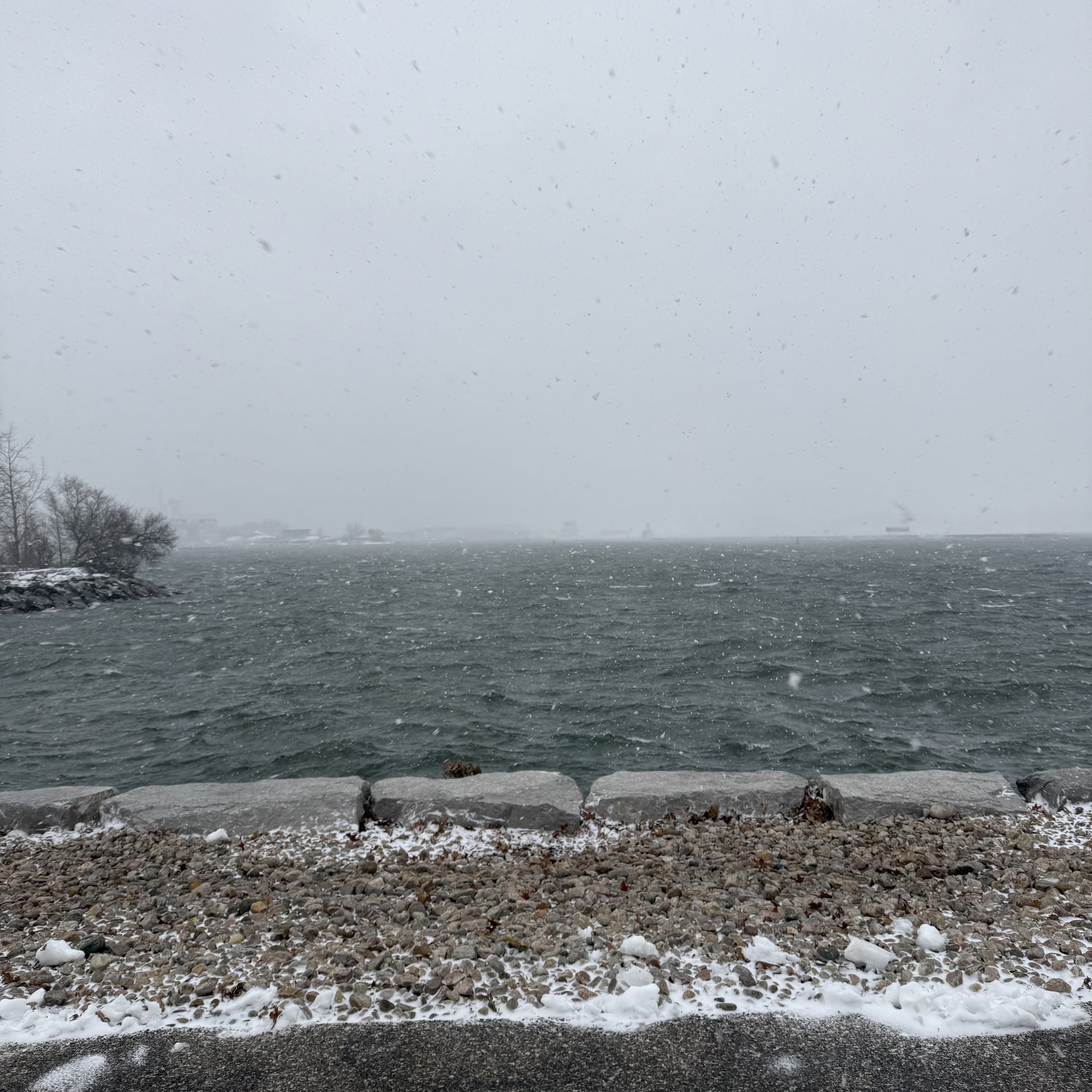 Pebble shoreline and concrete blocks at the water’s edge, rough lake water and falling snow stretching into a grey horizon.