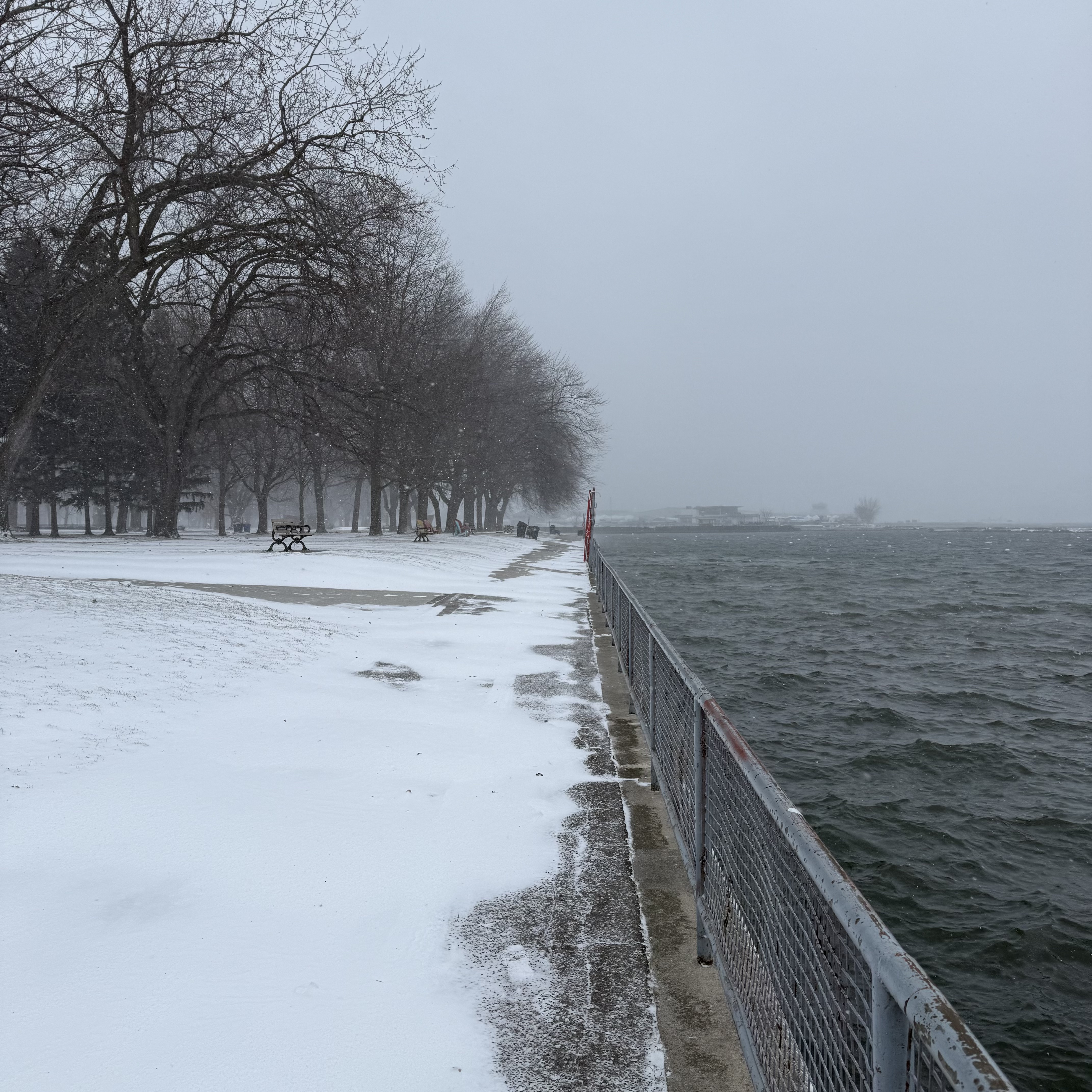 A long, snow-covered shoreline path lined with bare trees and benches, with a metal railing beside the choppy lake.
