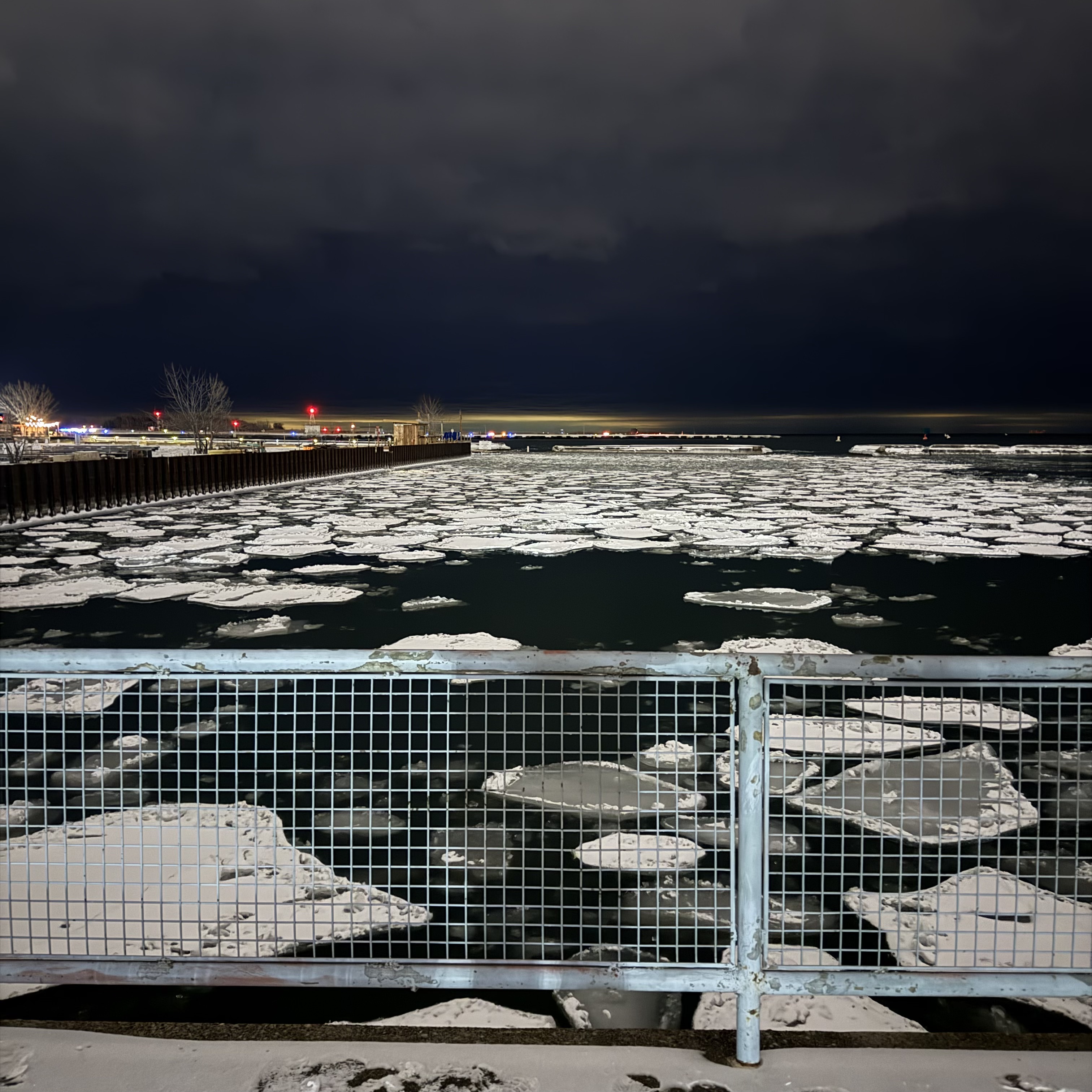 Nighttime view over dark water filled with broken sheets of floating ice, seen from behind a pale blue metal railing; distant red harbor lights glow under a low, stormy sky.