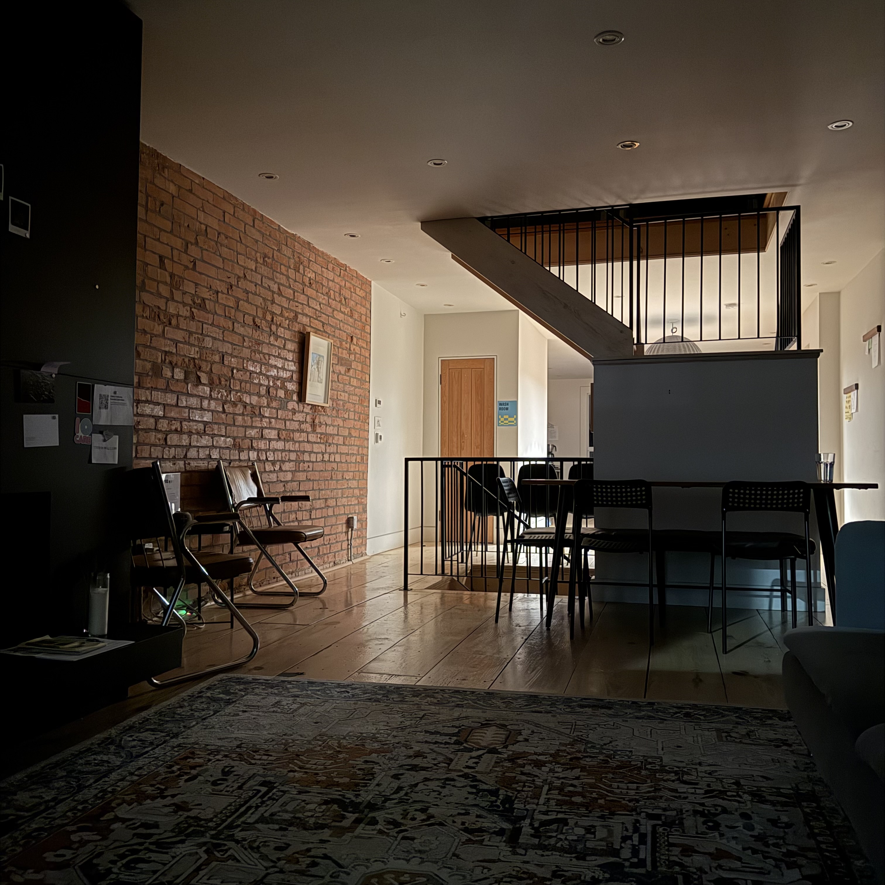 A dimly lit interior of a modern coworking or living space with a warm, minimal aesthetic. A brick accent wall runs along the left side, with two metal-framed chairs and a small bench placed against it. In the center is a dining table surrounded by simple black chairs, partially tucked under the landing of a staircase with black railings. Light wood floors and a muted, patterned rug fill the foreground. The room feels quiet, spacious, and slightly shadowed, as if lit only by natural light on an overcast day.