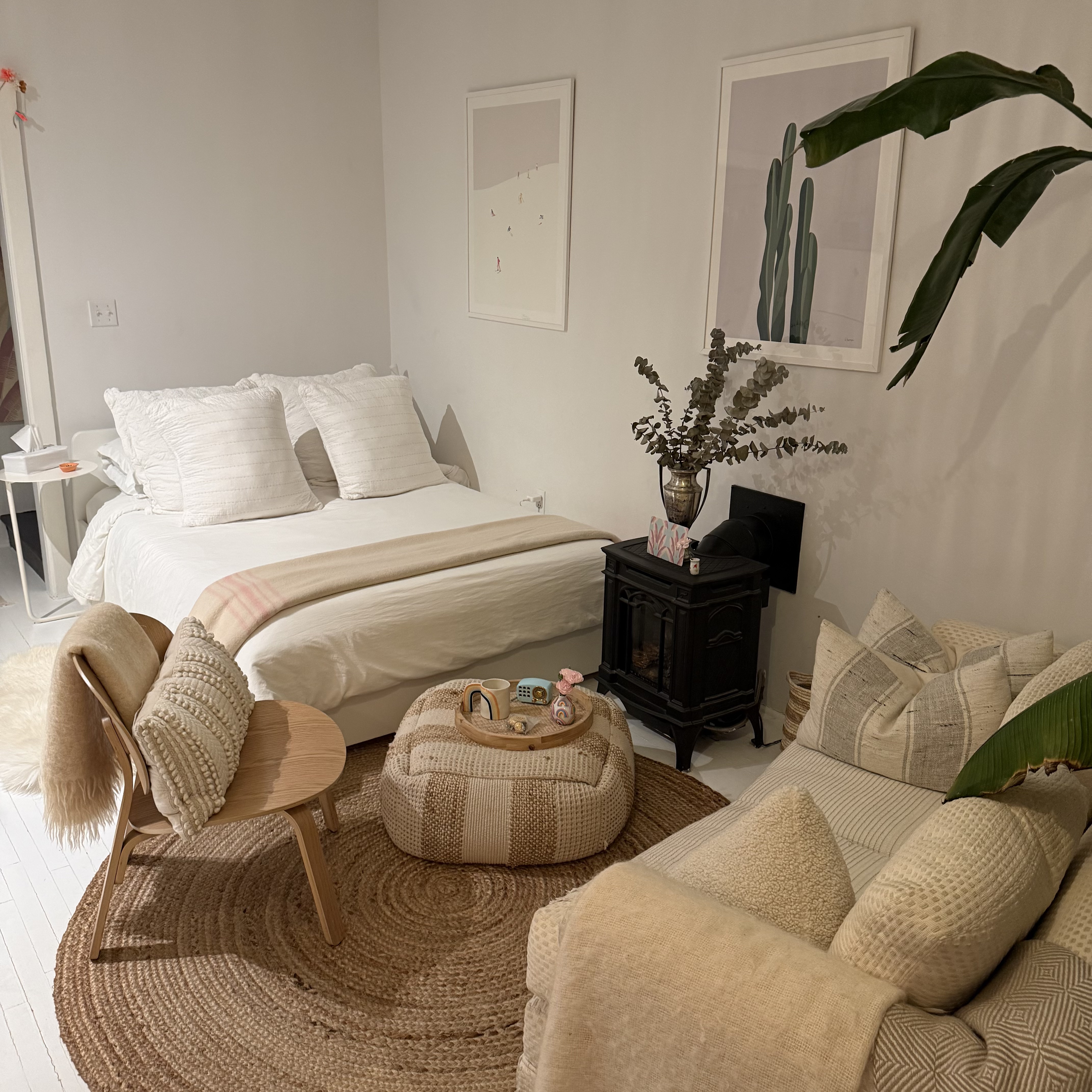 A cream sofa piled with linen pillows beside a bird of paradise plant and cast iron radiator, lit by a woven pendant lamp.