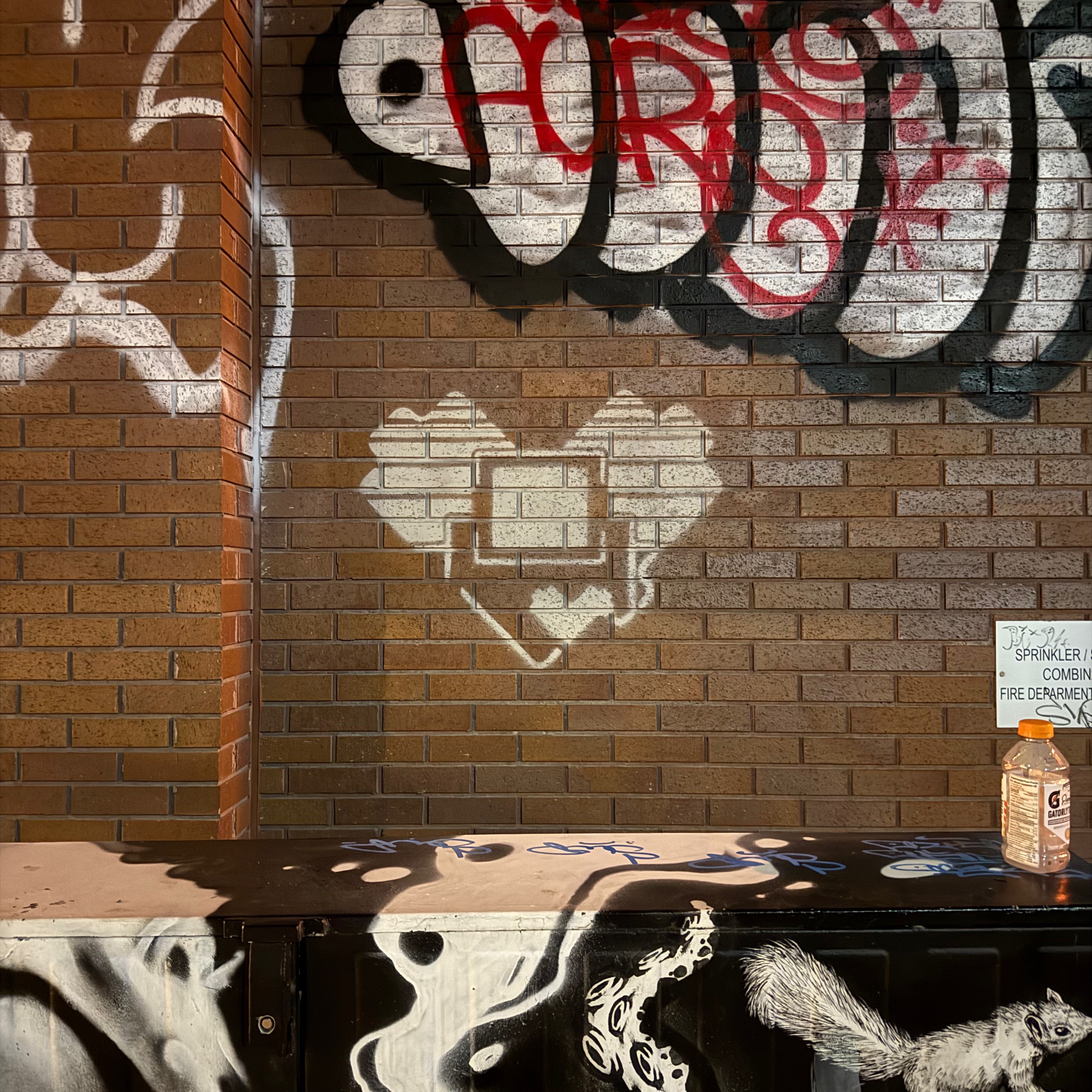 A brick wall covered in layered street art and graffiti. At the center, a white stencil of a small robot with a heart-shaped chest — the Toronto Lovebot — stands out sharply against the bricks. Surrounding it are bold black, red, and white tags and murals, including part of a painted animal figure. A plastic bottle sits on a ledge beneath a small sign, giving the scene a mix of urban grit and character.