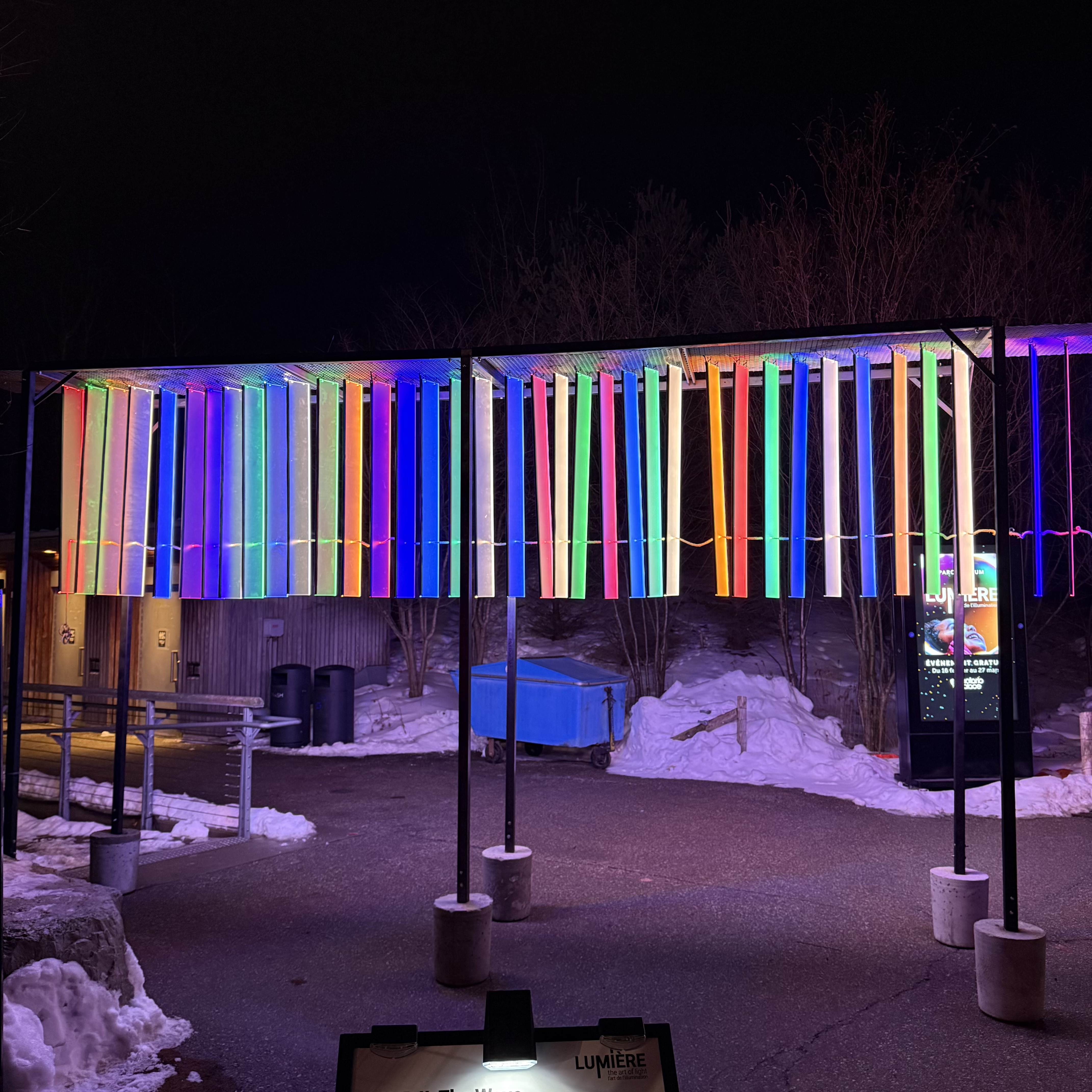 A canopy of vertical, translucent plastic light strips in vivid rainbow colours glows overhead at night, suspended from a black metal frame above snow and wet pavement in Trillium Park.
