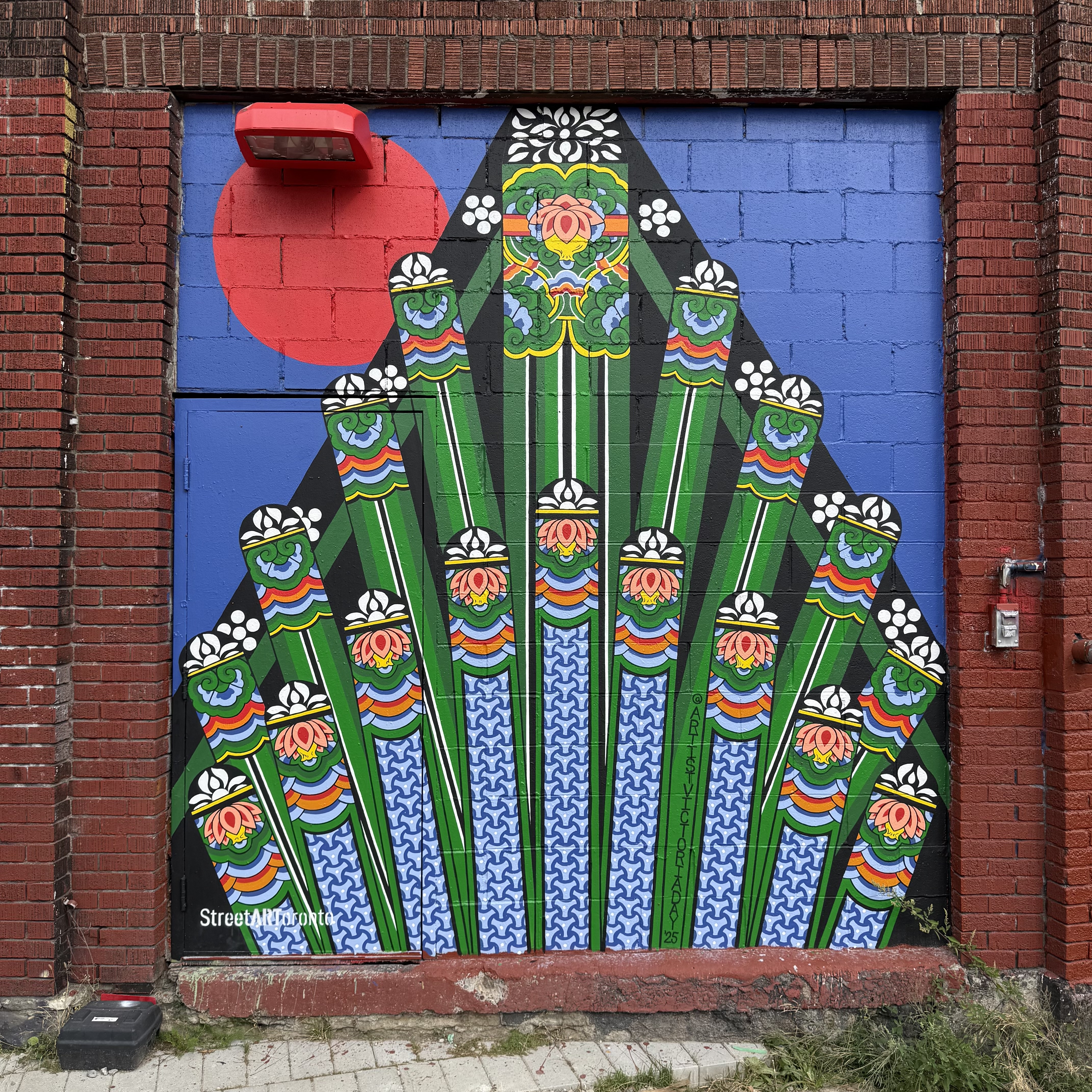 A bright, symmetrical mural painted on a brick wall, featuring a fan-like pattern of tall green columns decorated with colorful, floral designs and wave-like blue details. The background is split between deep blue and a vivid red circle in the top left, framed by dark red bricks. The geometric precision and bold palette give it a ceremonial, almost architectural feel.