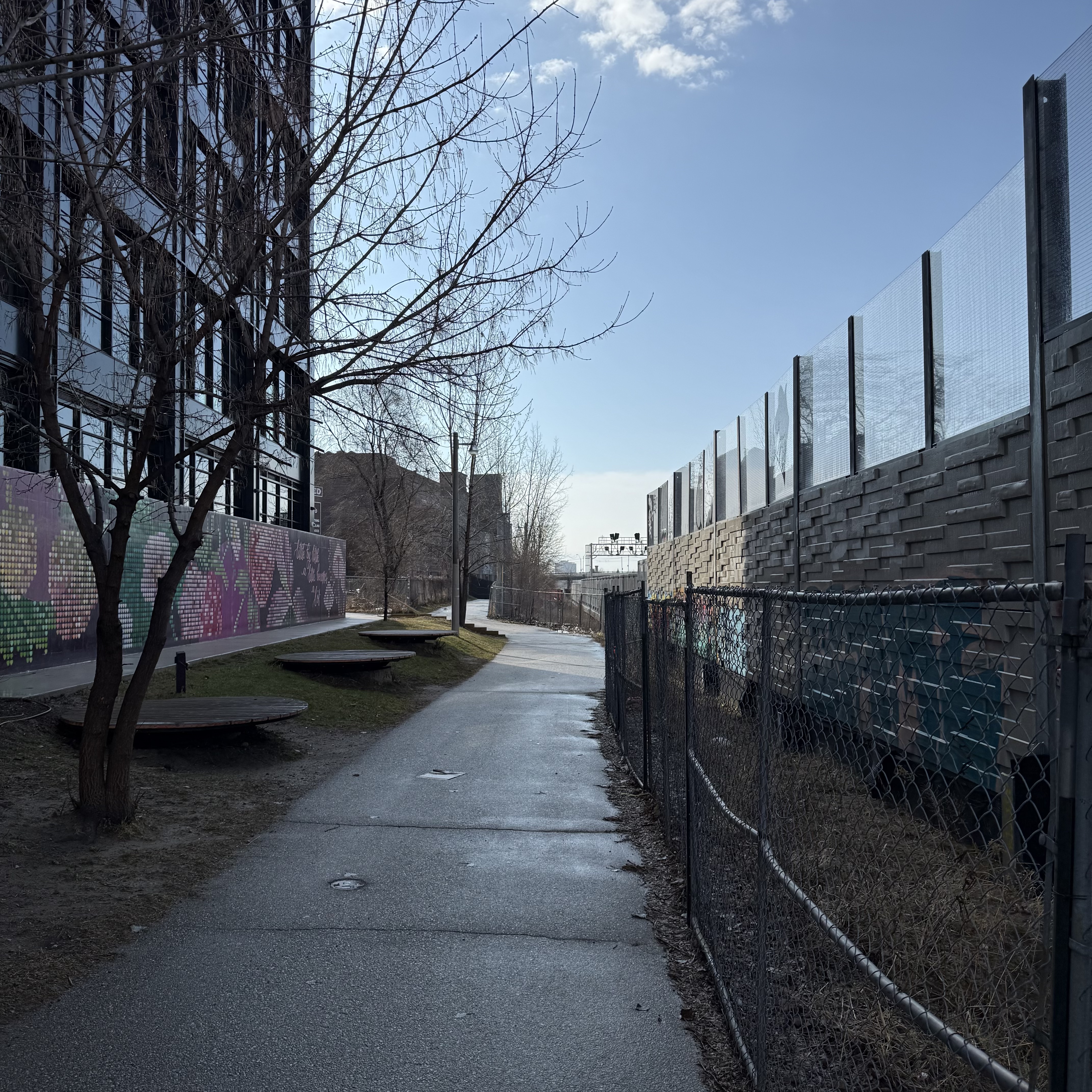 Paved path between a pixelated mural and a chain-link fence, with a concrete noise wall topped with glass panels on the right and a condo tower visible left. Bare trees, early spring.