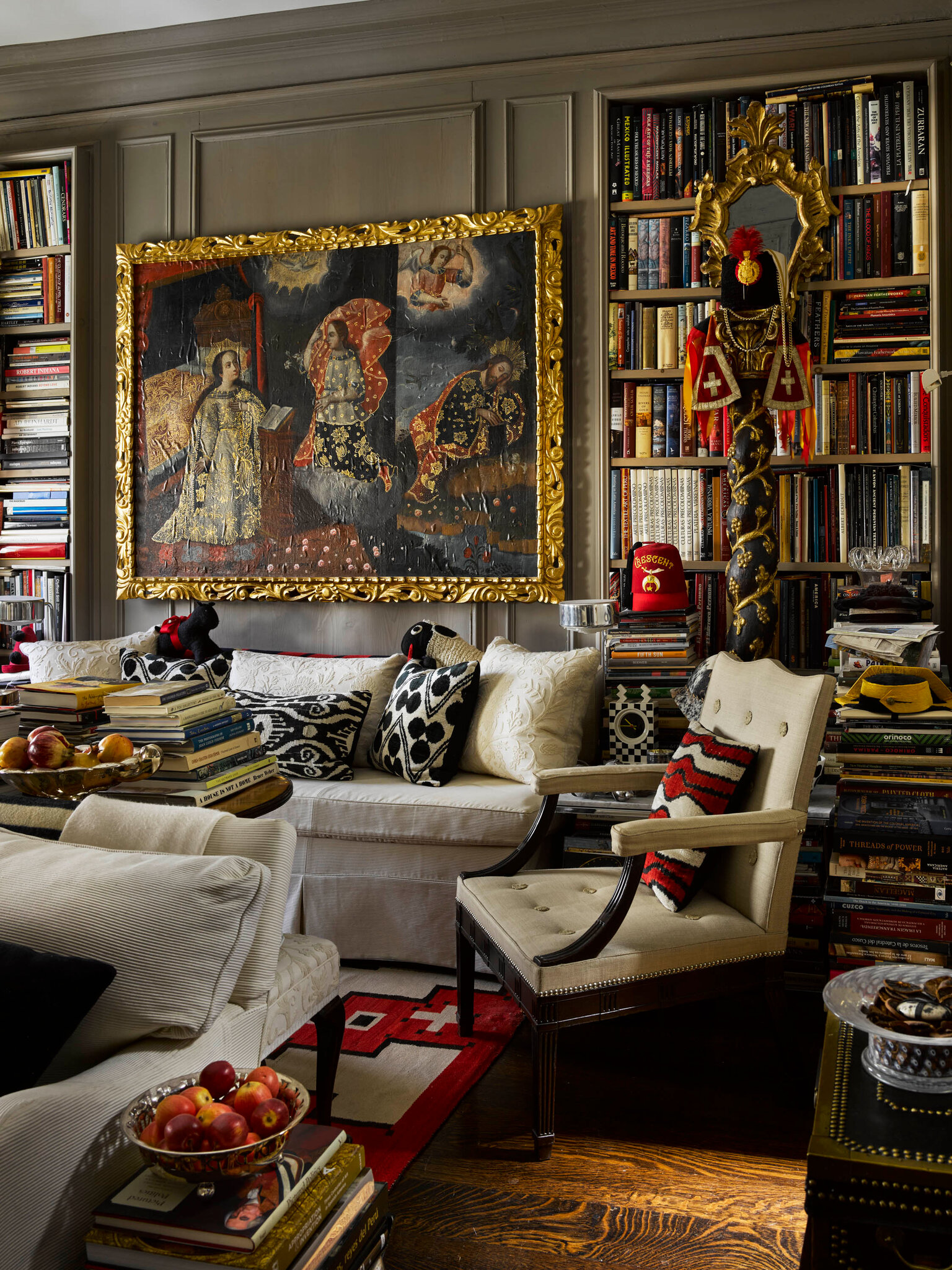 A richly layered sitting room lined floor-to-ceiling with bookshelves. A large gilded painting hangs over a white fireplace filled with logs, flanked by sconces and decorative ceramic figures. Stacks of books surround cream-coloured armchairs, a glass coffee table, and bowls of apples. The room feels warm, dense, and lived-in, with patterned rugs and abundant artwork.