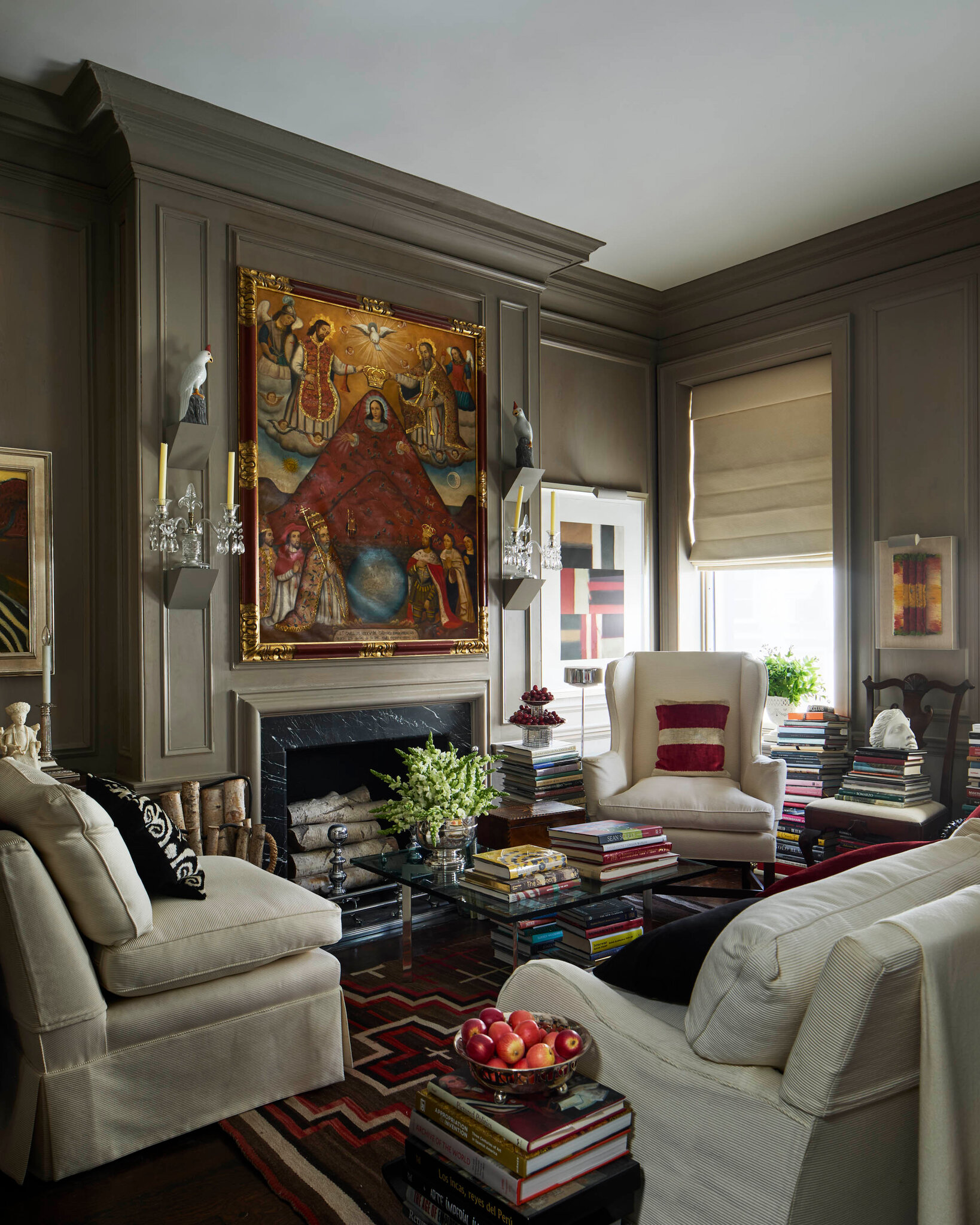 A cozy sitting room with taupe wall panelling and shelves overflowing with books. A monumental religious painting in a gold frame dominates the wall above a black fireplace. Cream armchairs encircle a glass coffee table perched on stacks of books. More piles of books line the window and flank every seat, creating a snug, library-like atmosphere.