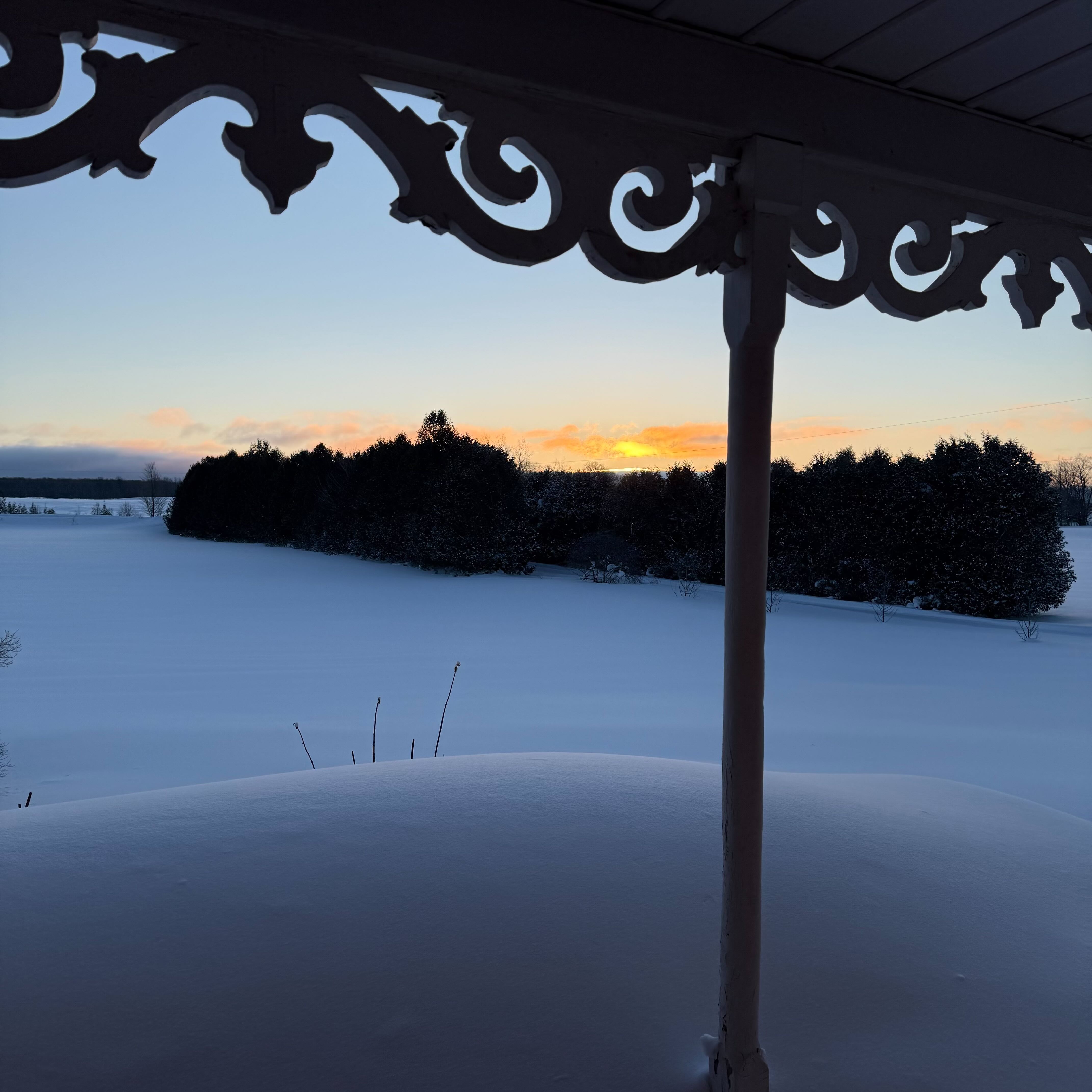 View from a covered porch overlooking a snow-covered field, with decorative trim framing a glowing sunrise behind a line of trees.