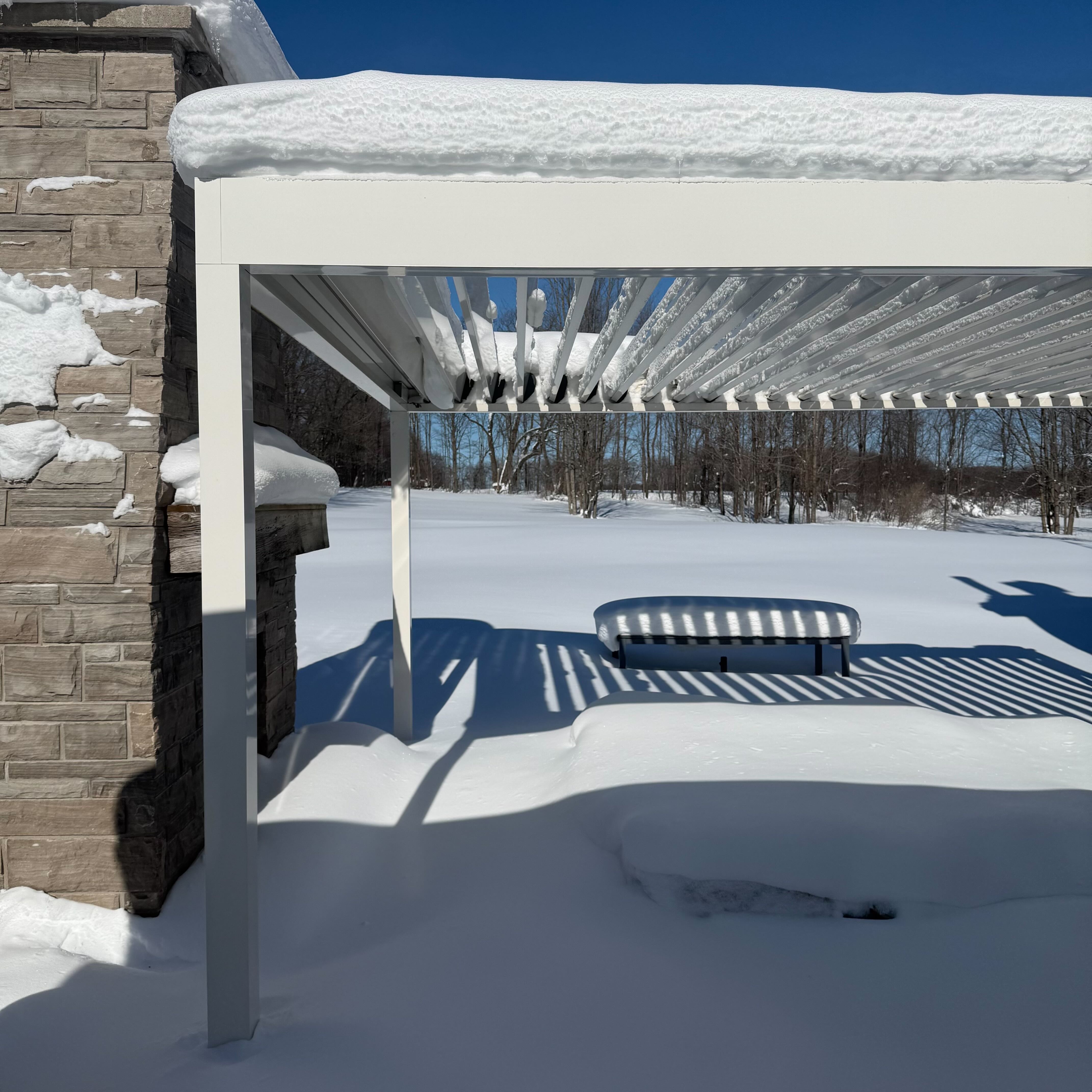 A modern pergola and patio buried under deep snow, casting striped shadows across the ground under a bright blue winter sky.