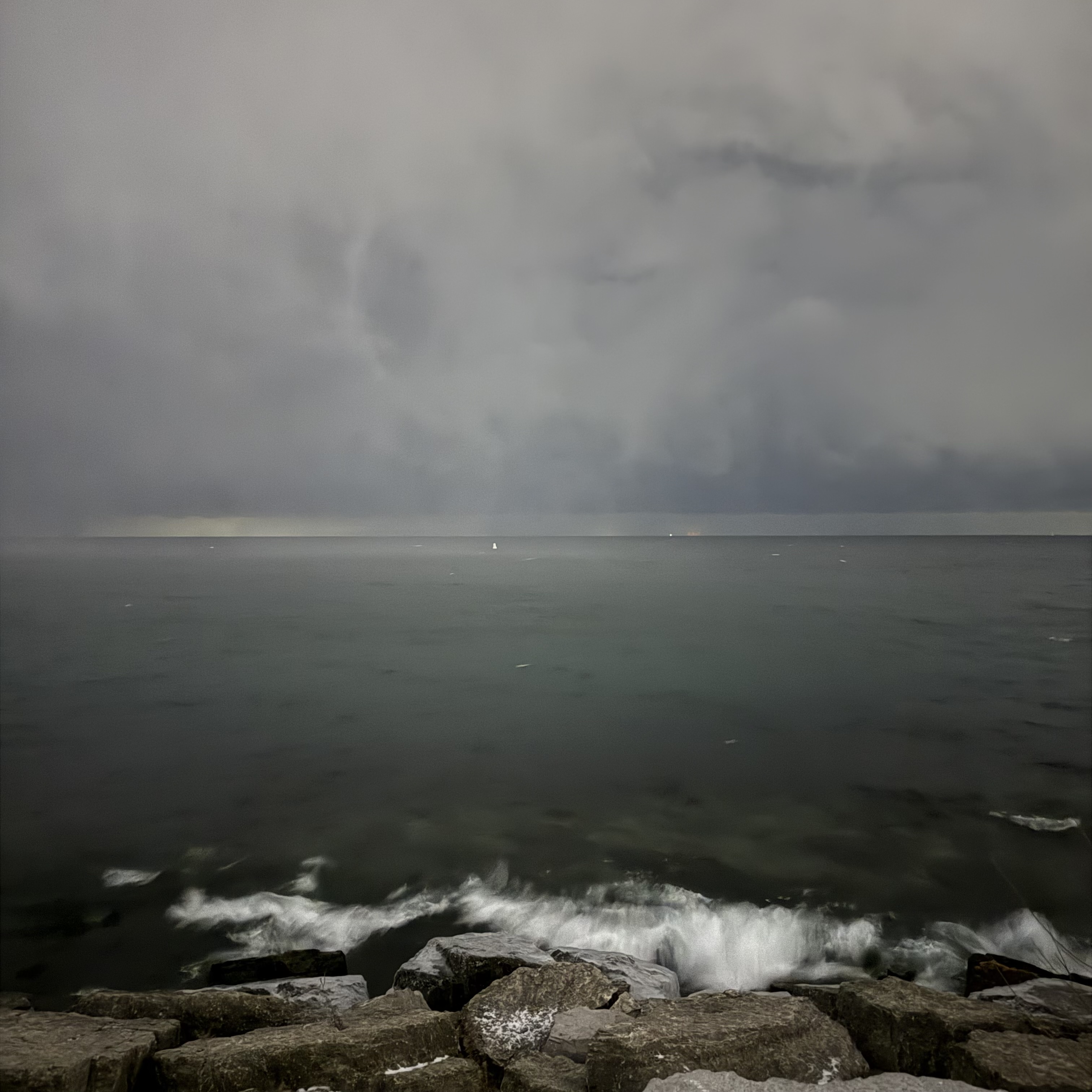 Dark, stormy view of Lake Ontario at night from a rocky shoreline, with small waves crashing against the rocks in the foreground and a low, grey horizon blending into a cloudy sky.