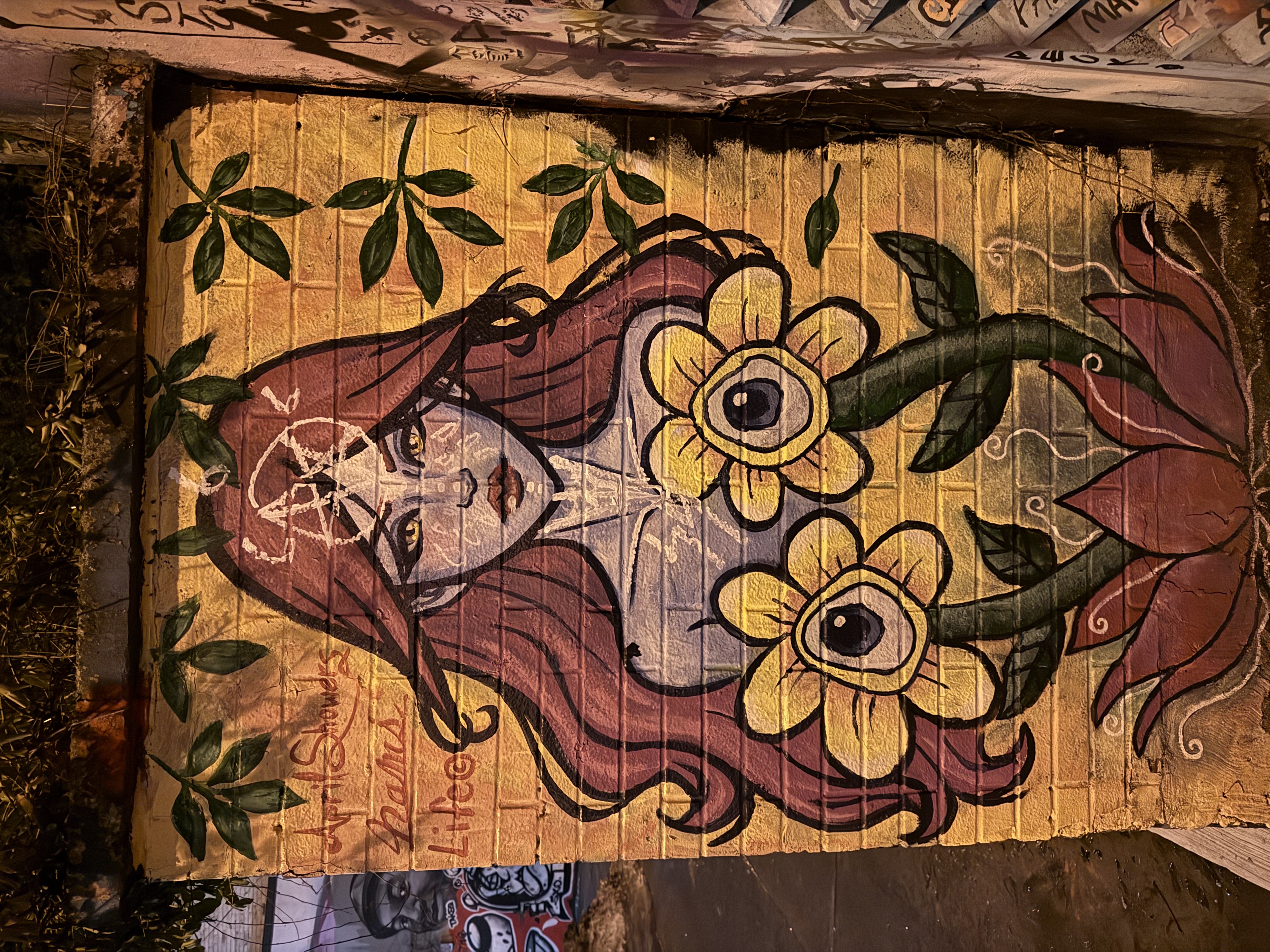 A street art mural on a yellow brick wall at night. A woman with long auburn hair and lavender skin holds two large yellow flowers with eyes at their centres. A pentagram marks her forehead; green vines frame the scene.