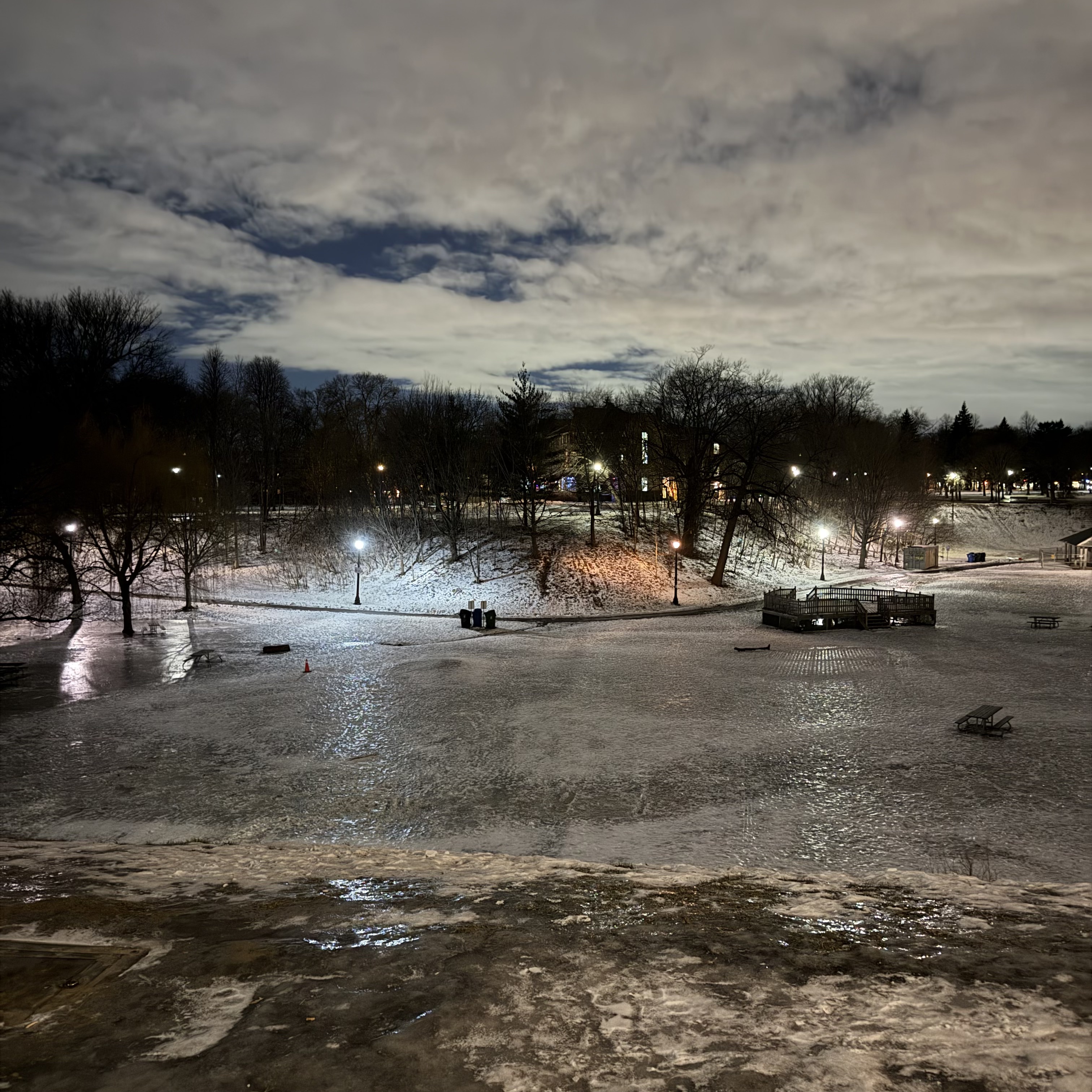 Night view over a frozen, glossy pond in a snowy park, with bare trees silhouetted against a cloudy sky and a line of lampposts lighting the path and a small wooden platform on the ice.