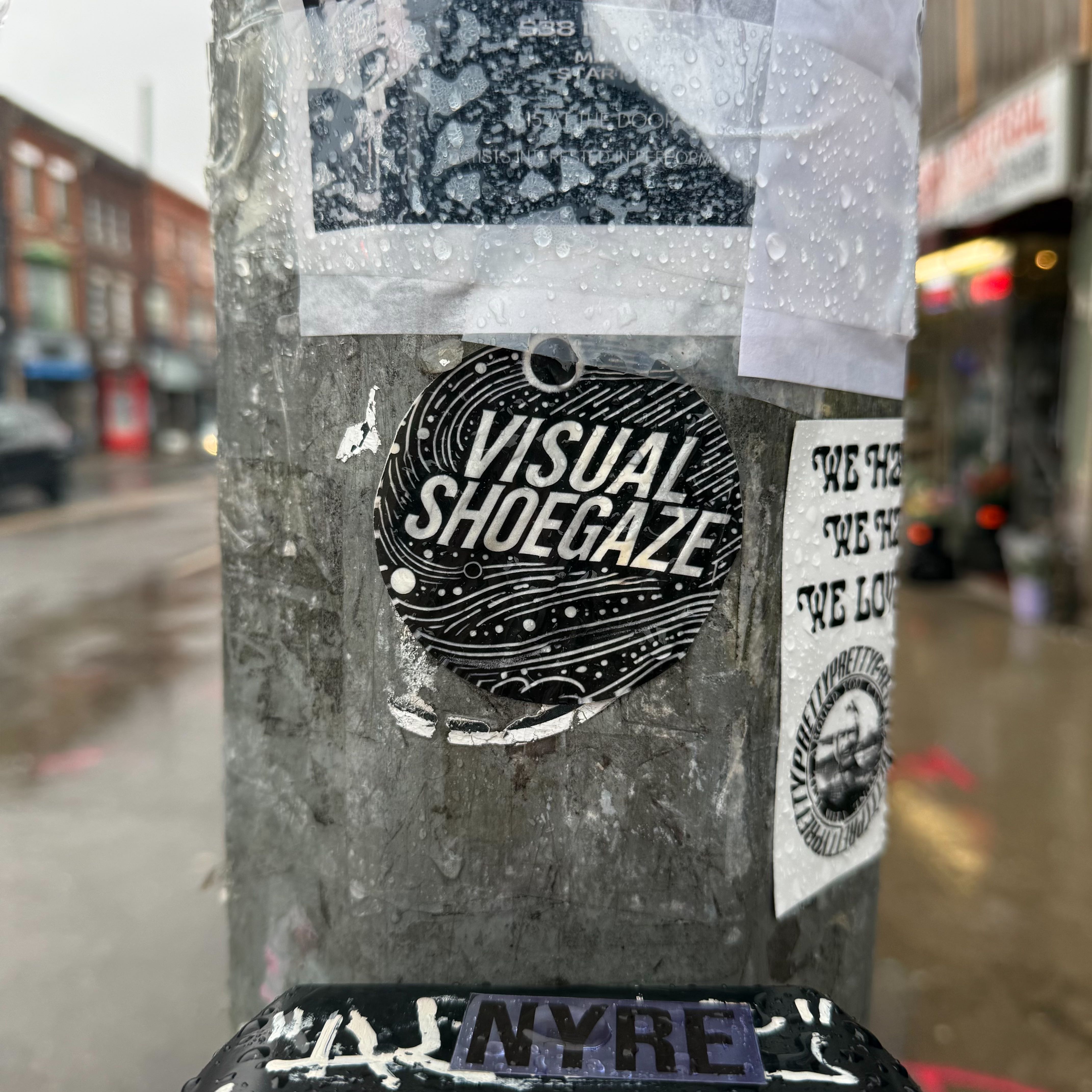 A black circular sticker reading 'Visual Shoegaze' in bold white lettering, mounted on a weathered concrete pole on Ossington Ave, surrounded by other stickers.