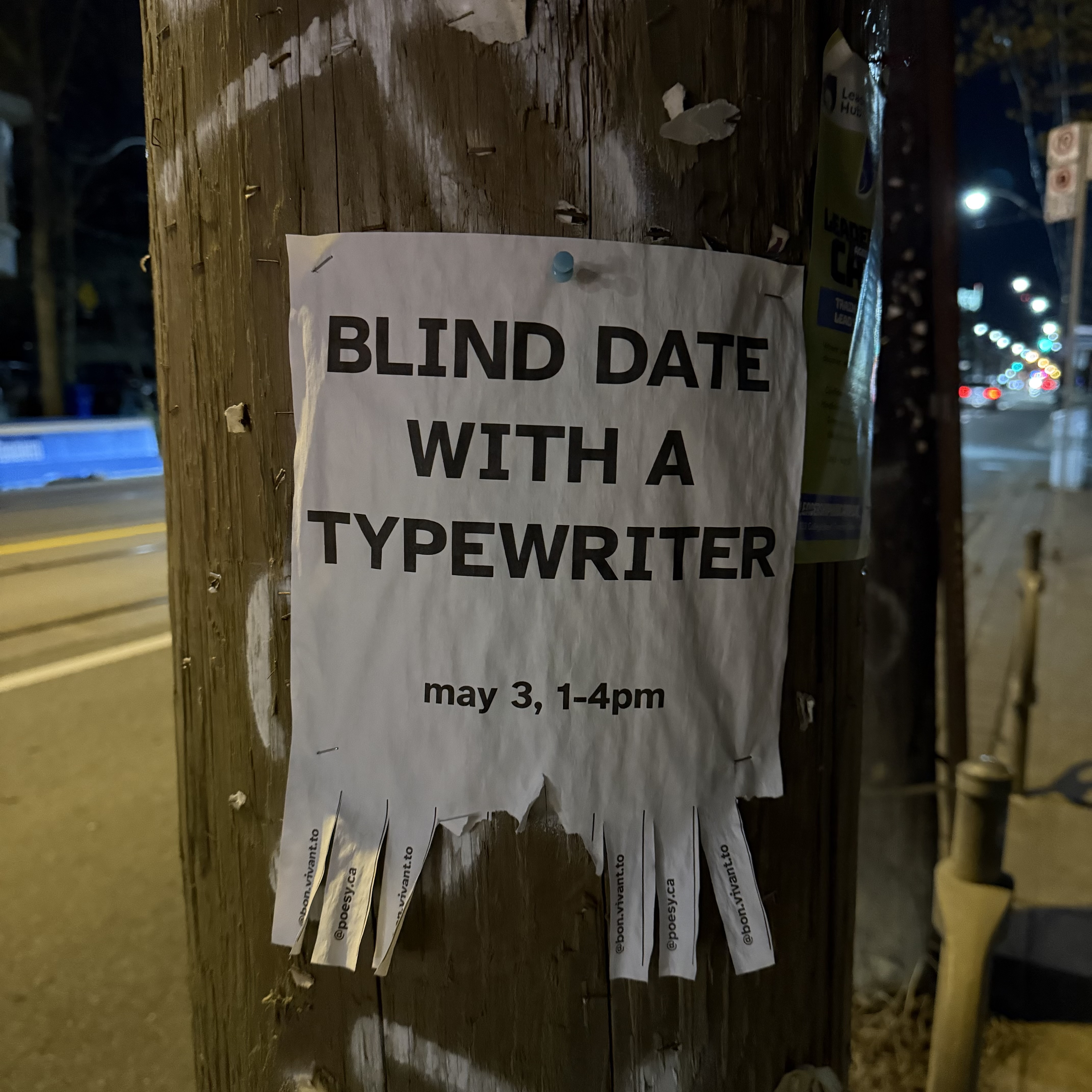 A white flyer pinned with a teal thumbtack to a weathered wooden utility pole at night. Bold black caps read "BLIND DATE WITH A TYPEWRITER" above smaller lowercase "may 3, 1–4pm". Fringed tear-off strips along the bottom carry the handles @bon.vivant.to and @poesy.ca, a few already pulled. The pole is scarred with staples and old paper scraps; behind it, a dark street recedes into blurred streetlights.