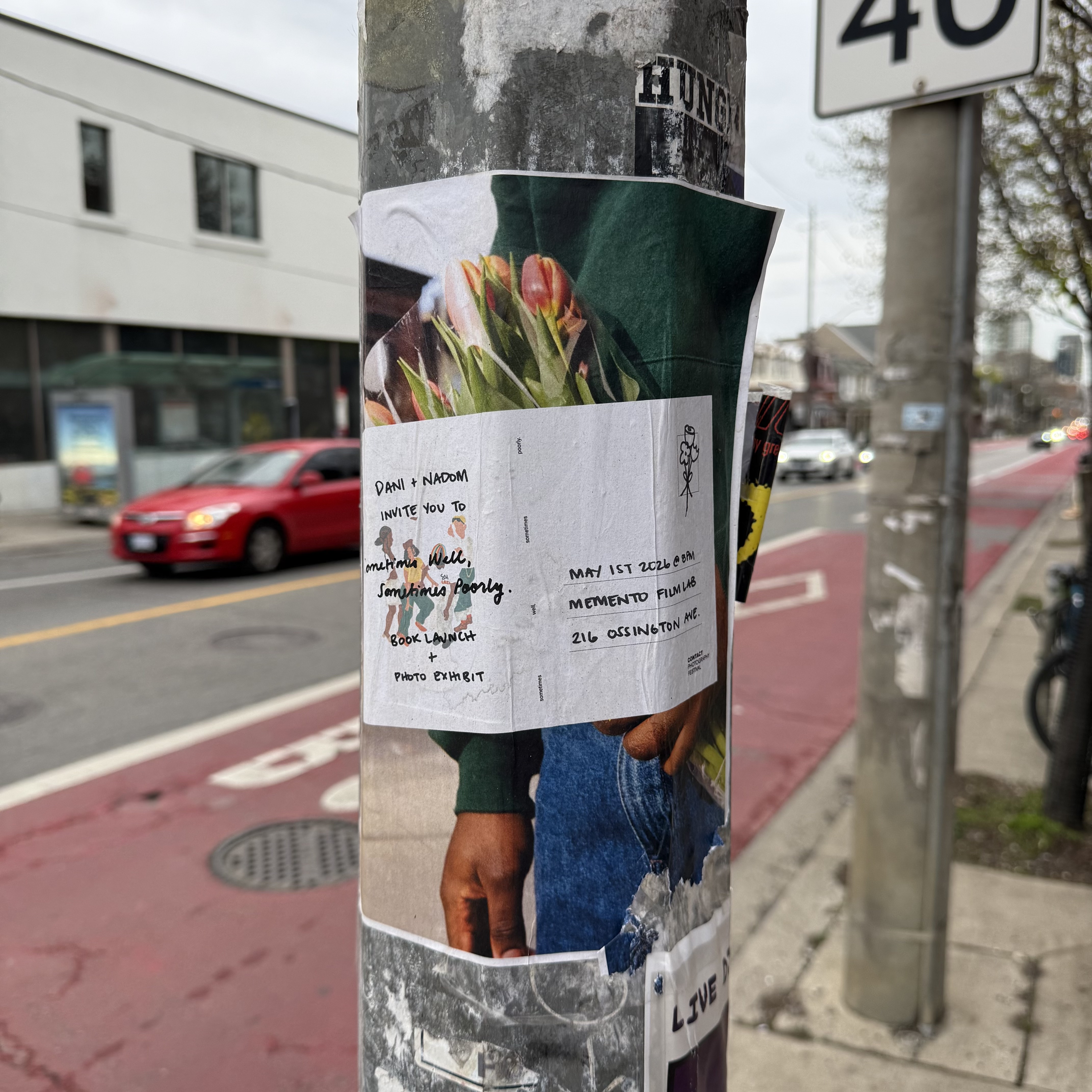 A wheat-pasted poster on a utility pole on a city street. The poster layers a photograph of someone in a green hoodie holding a bouquet of pink tulips over a white event flyer, which reads: "Dani + Nadom invite you to Sometimes Well, Sometimes Poorly. Book Launch + Photo Exhibit. May 1st 2026 @ 8PM. Memento Film Lab, 216 Ossington Ave." A 40km/h speed sign hangs above. Behind the pole: a red bike lane, passing cars, and a grey spring sky.
